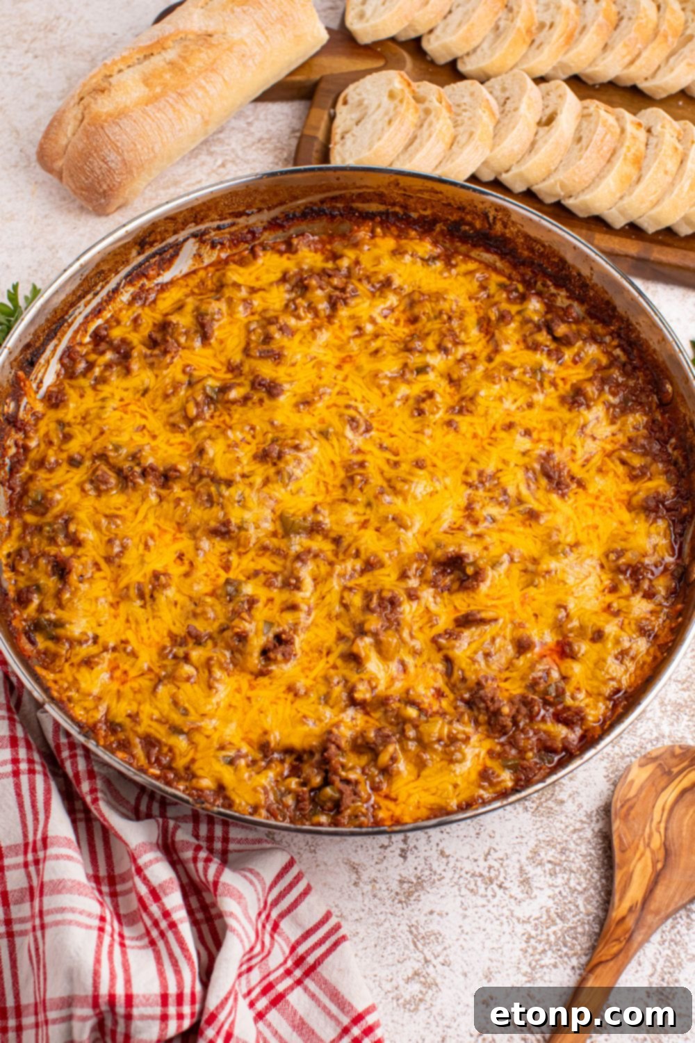 Ultimate Sloppy Joe Dip 5 A large pan of hot Sloppy Joe Dip, with a rich, savory surface, placed on a wooden table alongside a cutting board with neatly sliced bread.