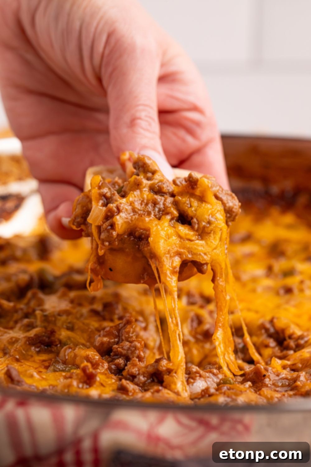 Ultimate Sloppy Joe Dip 7 A person's hand with a wooden spoon digging into a pan of savory Sloppy Joe Dip, ready to enjoy a hearty scoop.
