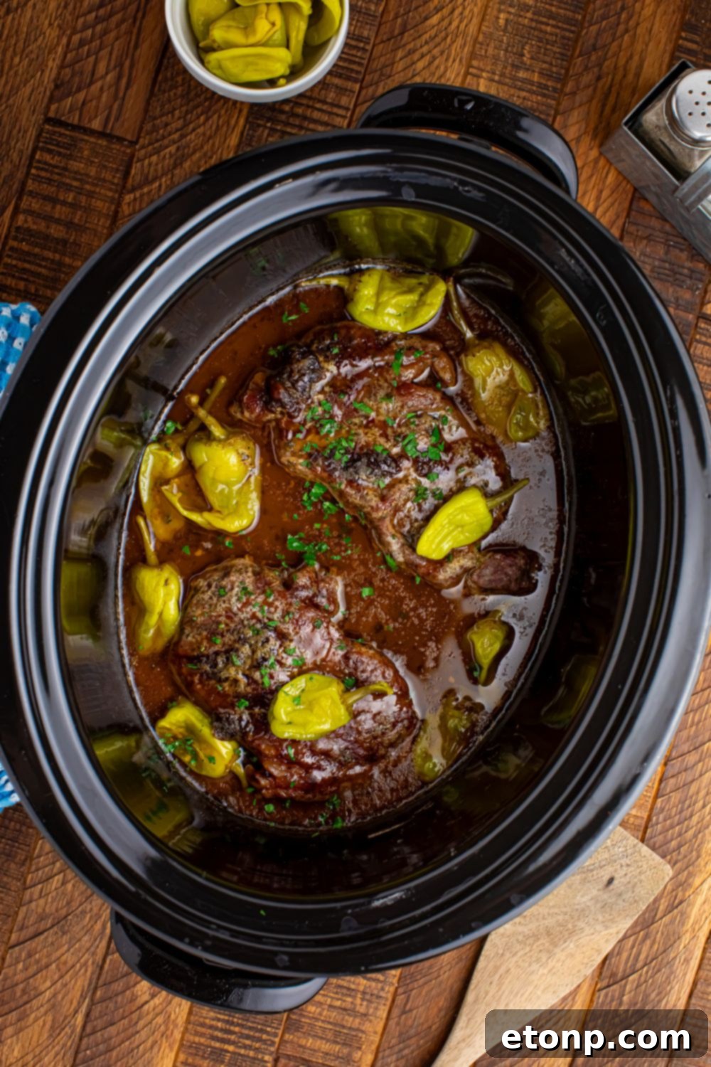Slow Cooker Mississippi Ribeye Steaks in crock po on wooden table wih peppers in a bowl tto the side