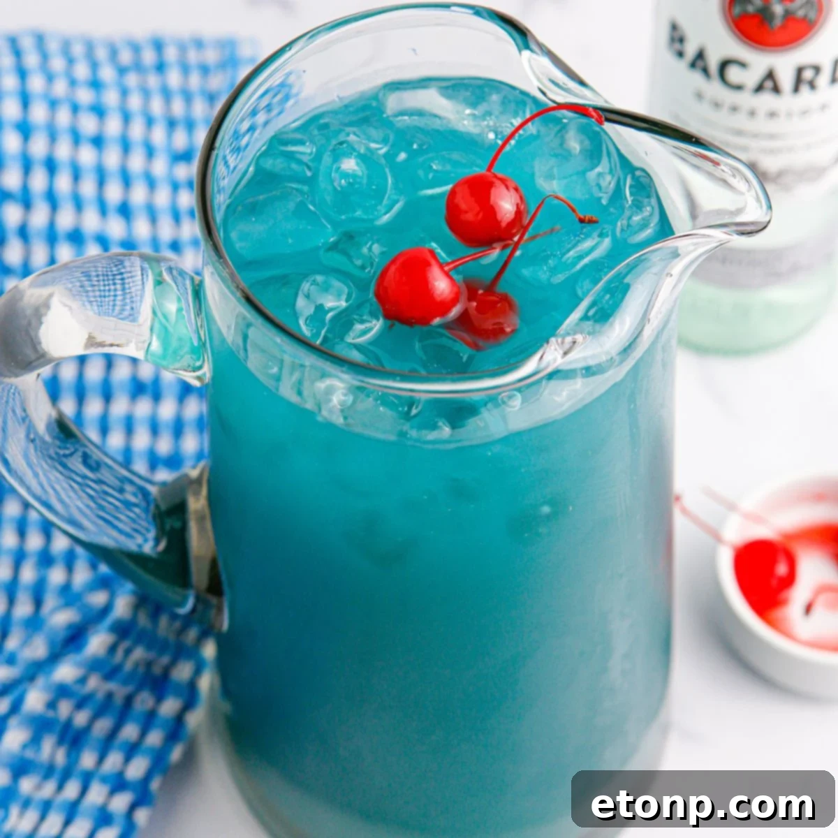 Blue Hawaiian Punch in a pitcher on table with a bowl of cherries on side and a bottle of Bacardi in the background