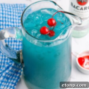 Boozy Blue Hawaiian Punch in a pitcher on table with a bowl of cherries and a bottle of Bacardi