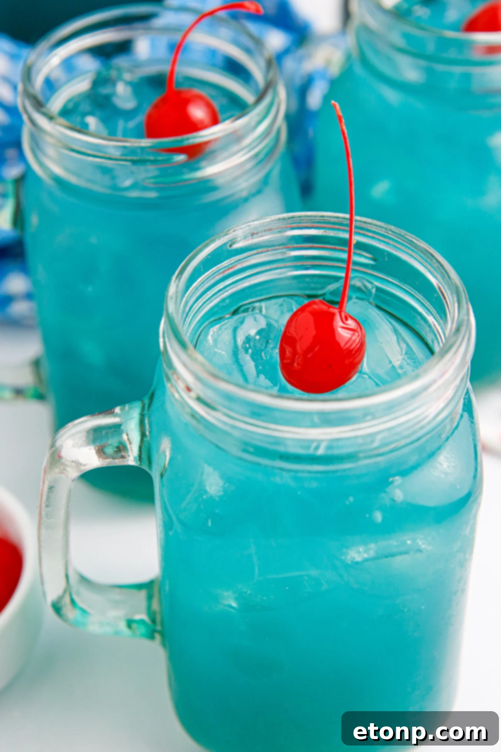 Two glasses of Blue Hawaiian Punch on a table, one with a straw and cherry