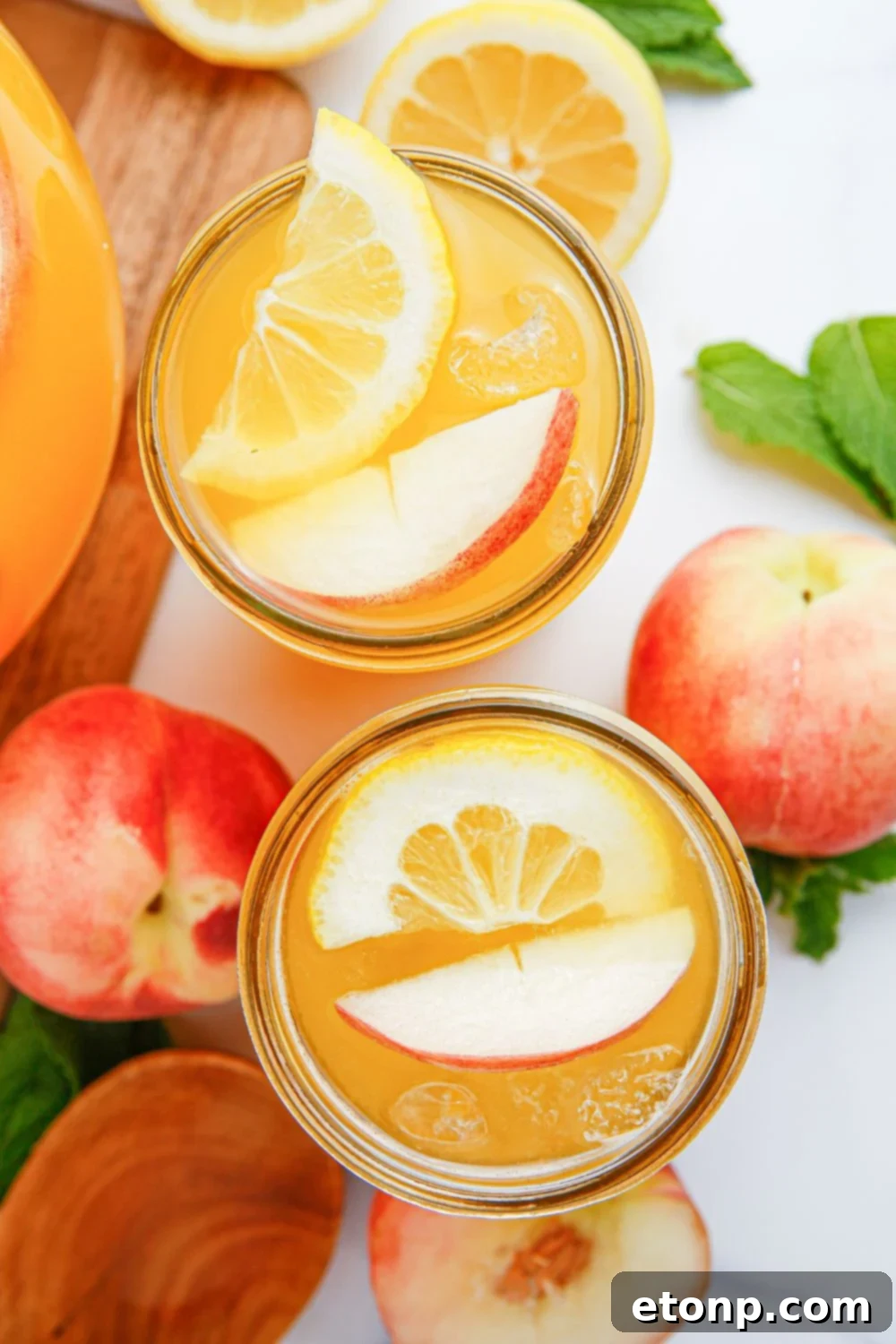 Glasses of peach lemonade are elegantly arranged on a table, accompanied by slices of fresh peaches and lemons, along with vibrant mint leaves, creating a visually appealing and utterly refreshing scene.
