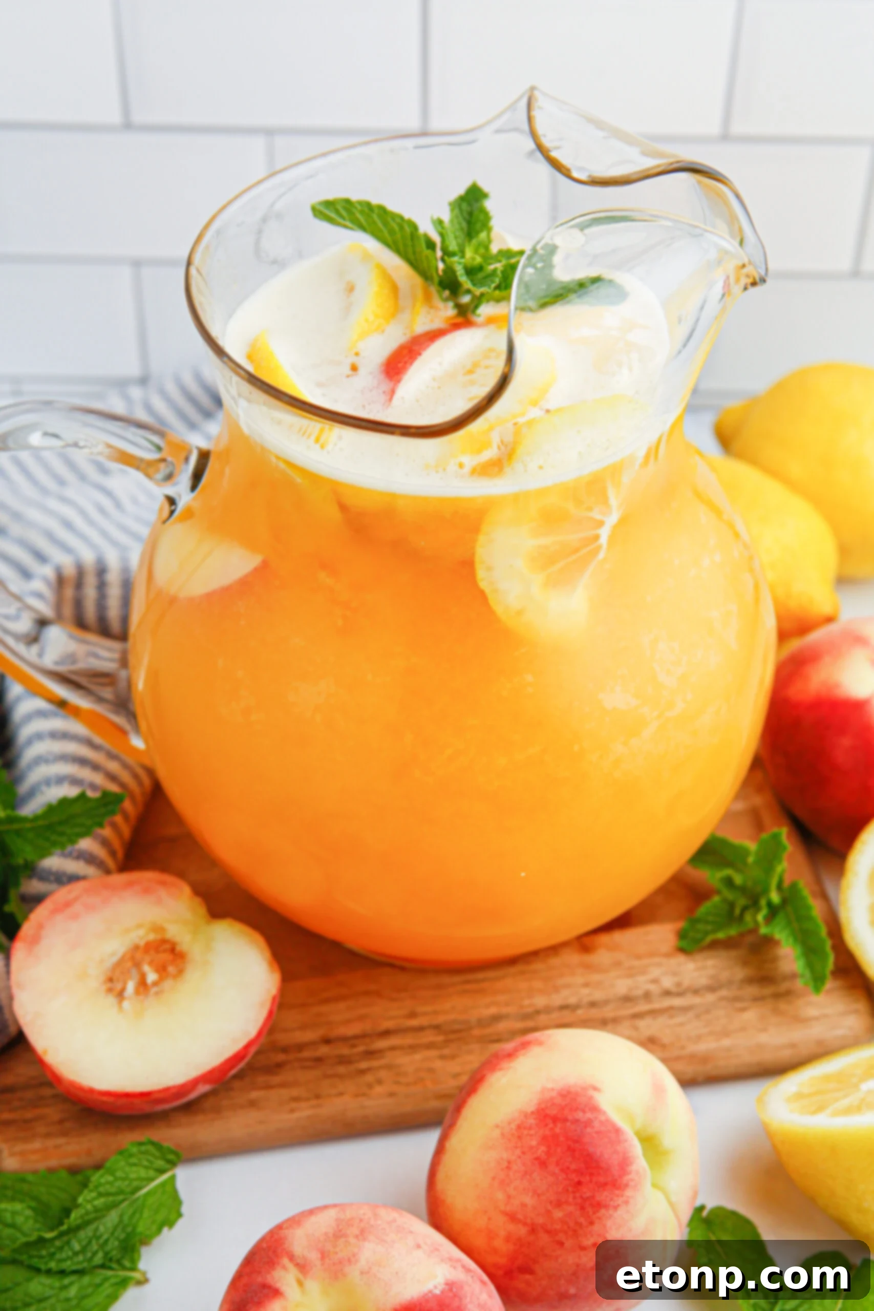 A clear pitcher of peach lemonade, elegantly garnished with a peach slice and orange slices, sits beautifully surrounded by fresh peaches, creating an inviting and visually appealing summer drink display.