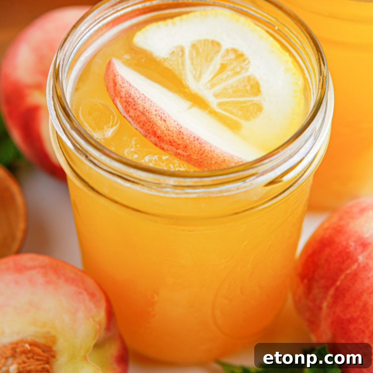 A vibrant glass of homemade Peach Lemonade, adorned with a fresh peach slice and a bright orange slice. More plump, ripe peaches surround the rustic mason jar, highlighting the natural goodness within.