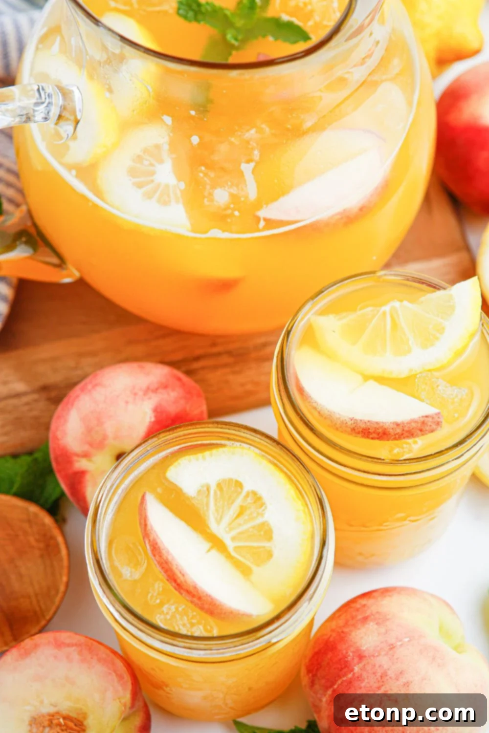 A large glass pitcher brimming with homemade peach lemonade sits invitingly on a table. Surrounding it are several mason jars, also full of the refreshing drink, along with scattered fresh peaches, ready to be enjoyed.