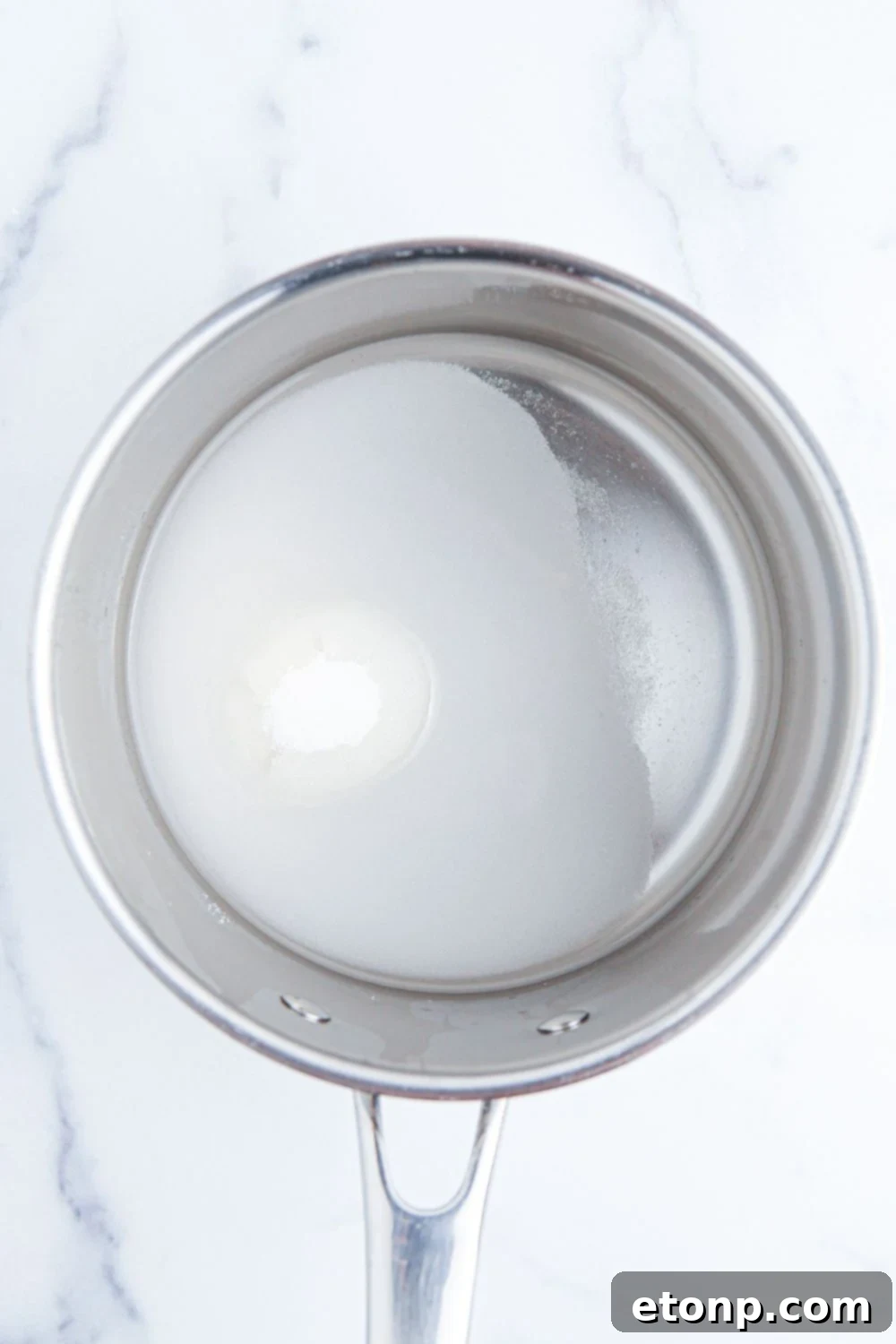 A small saucepan on a table contains a clear mixture of sugar and water, gently simmering to create a simple syrup, a key component for sweetening beverages.