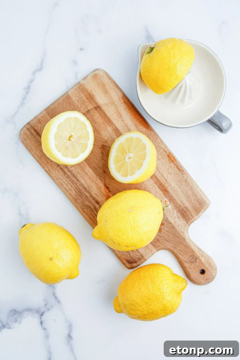 Fresh lemons, both whole and halved, are artfully arranged on a wooden cutting board. A citrus juicer sits ready next to a half-lemon, anticipating the extraction of vibrant lemon juice.