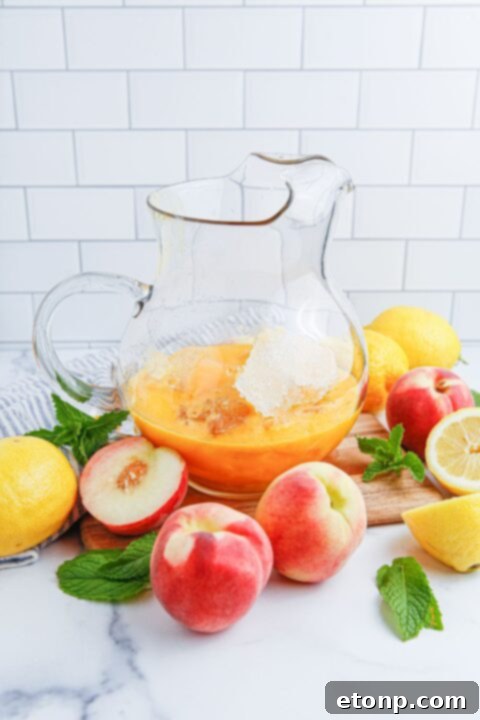 A large pitcher holds a concentrated peach mixture, with ice cubes and fresh peach and lemon slices surrounding it, showcasing the rich fruit essence before dilution into lemonade.