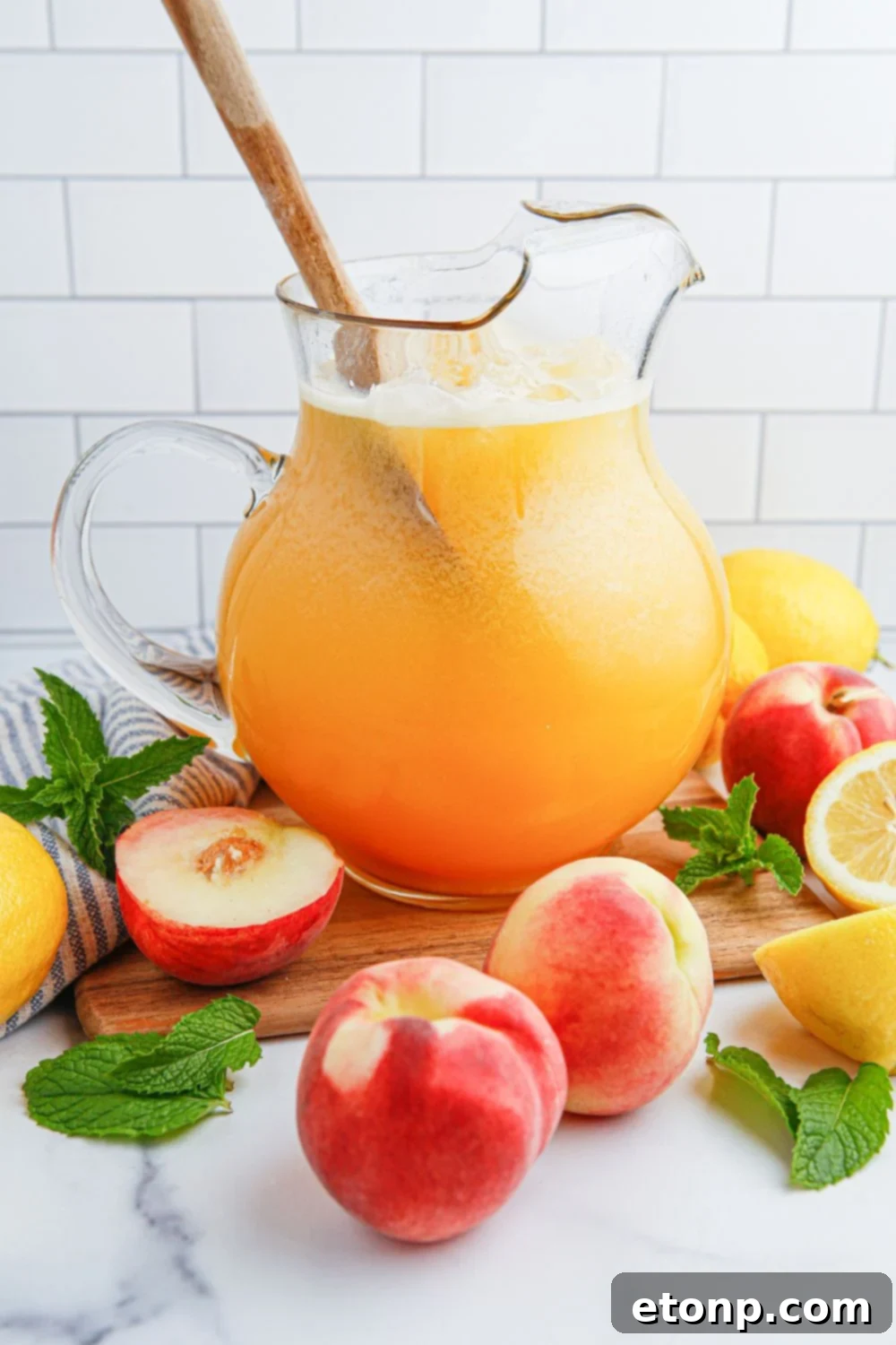 A wooden spoon stirs a pitcher of refreshing Peach Lemonade, while fresh sliced peaches and lemon wedges are elegantly arranged around the container, highlighting the natural ingredients and inviting a taste.
