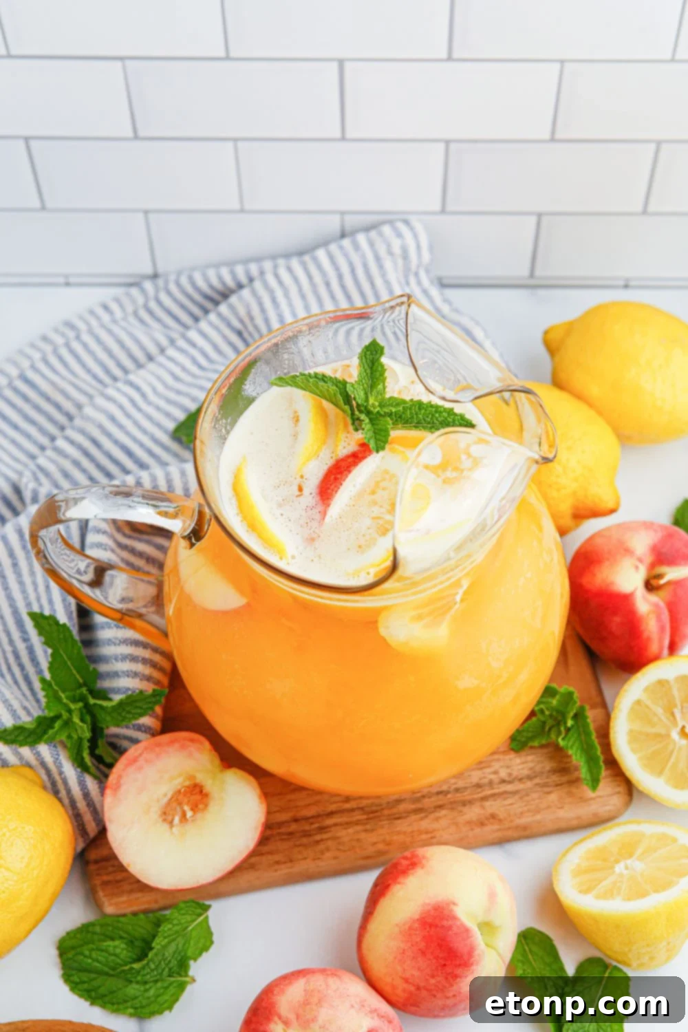 A pitcher of vibrant Peach Lemonade rests on a wooden cutting board, surrounded by an assortment of fresh peaches and lemons on a table, emphasizing the natural beauty and freshness of the ingredients.