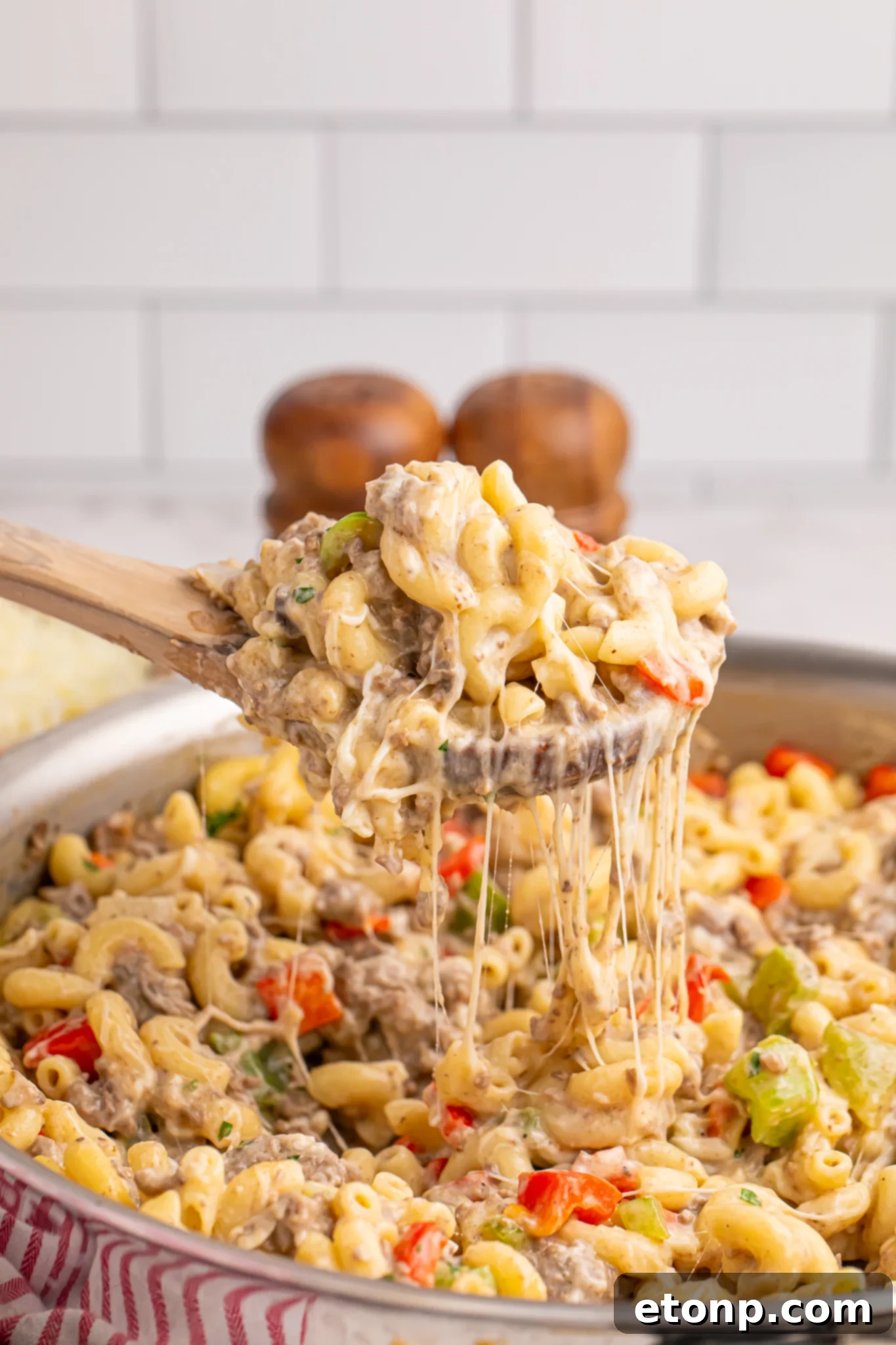 cheese steak pasta in a silver skillet with a wooden spoon lifting up a portion