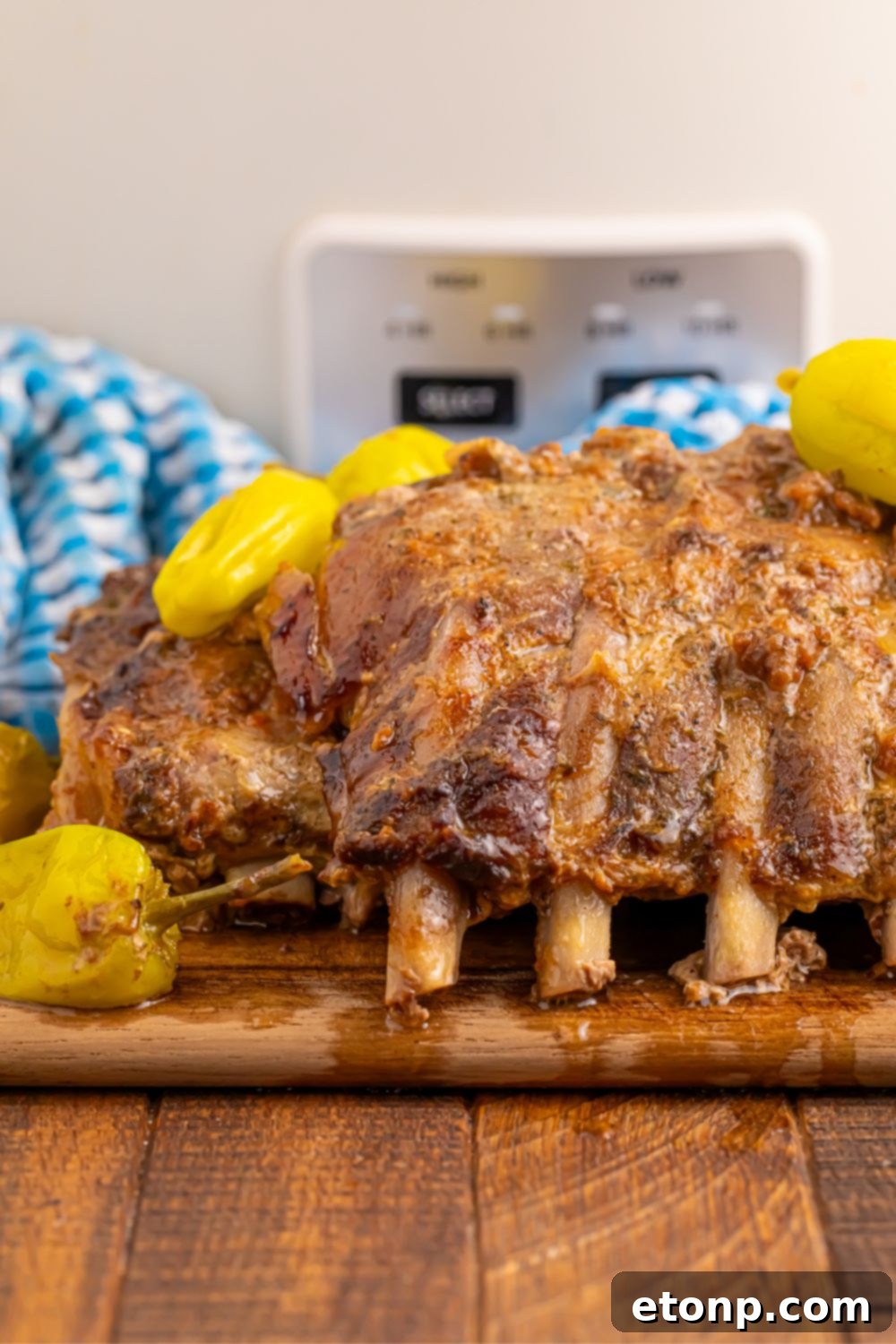 Cooked Slow Cooker Mississippi Ribs resting on a wooden cutting board, with a slow cooker and a blue towel in the softly blurred background.