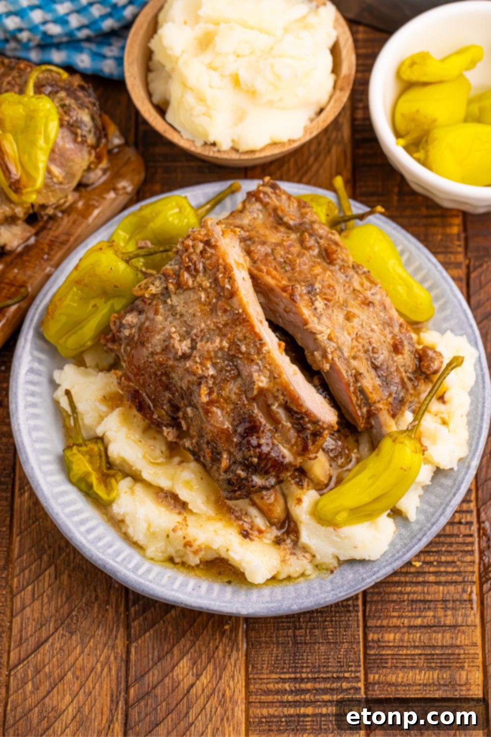 A full plate of Slow Cooker Mississippi Ribs alongside mashed potatoes and pepperoncini, with small bowls of individual ingredients blurred in the background, highlighting the meal's components.