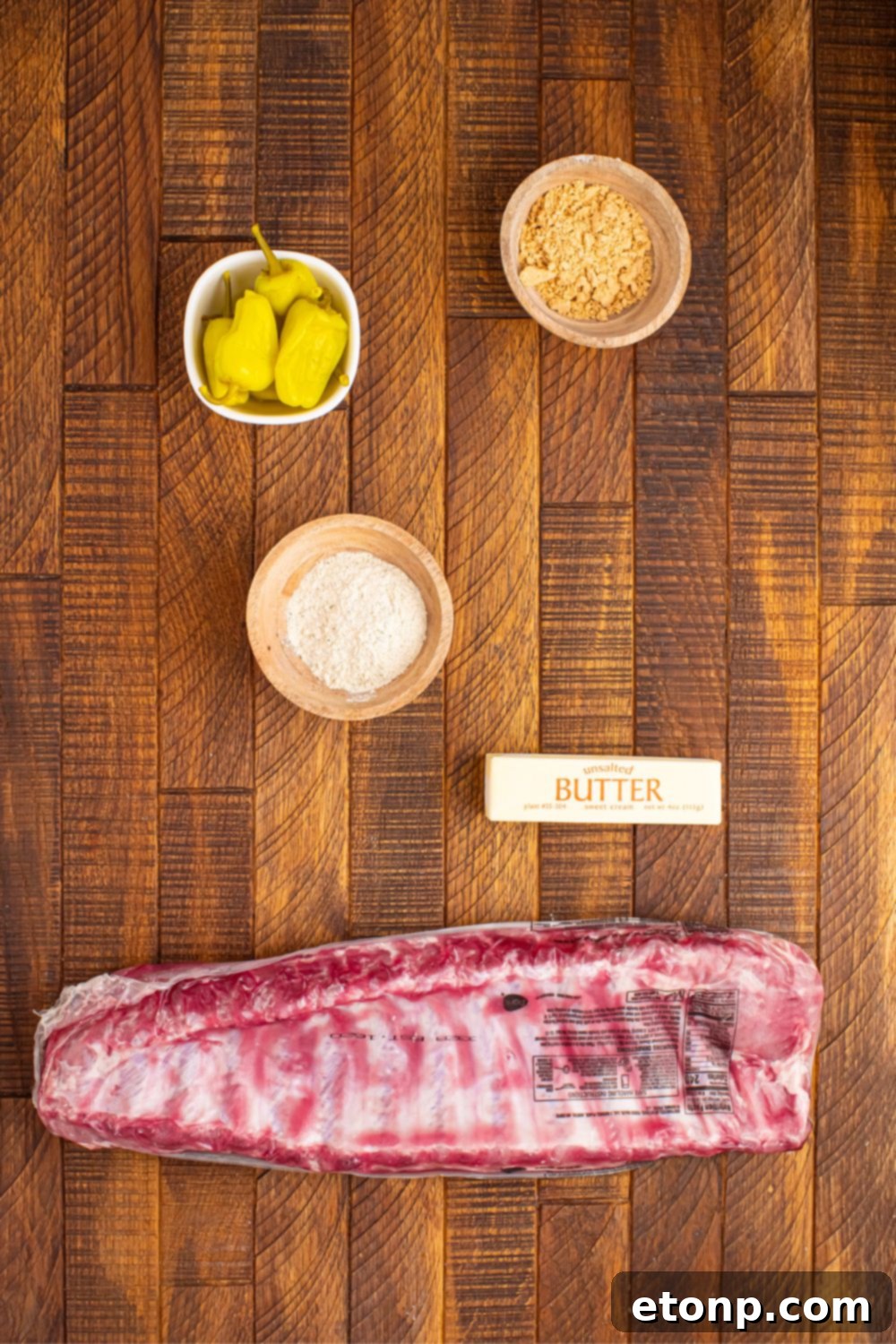 Individual ingredients for Slow Cooker Mississippi Ribs laid out: baby back ribs, a stick of butter, ranch seasoning packet, au jus seasoning packet, and a jar of pepperoncini peppers.