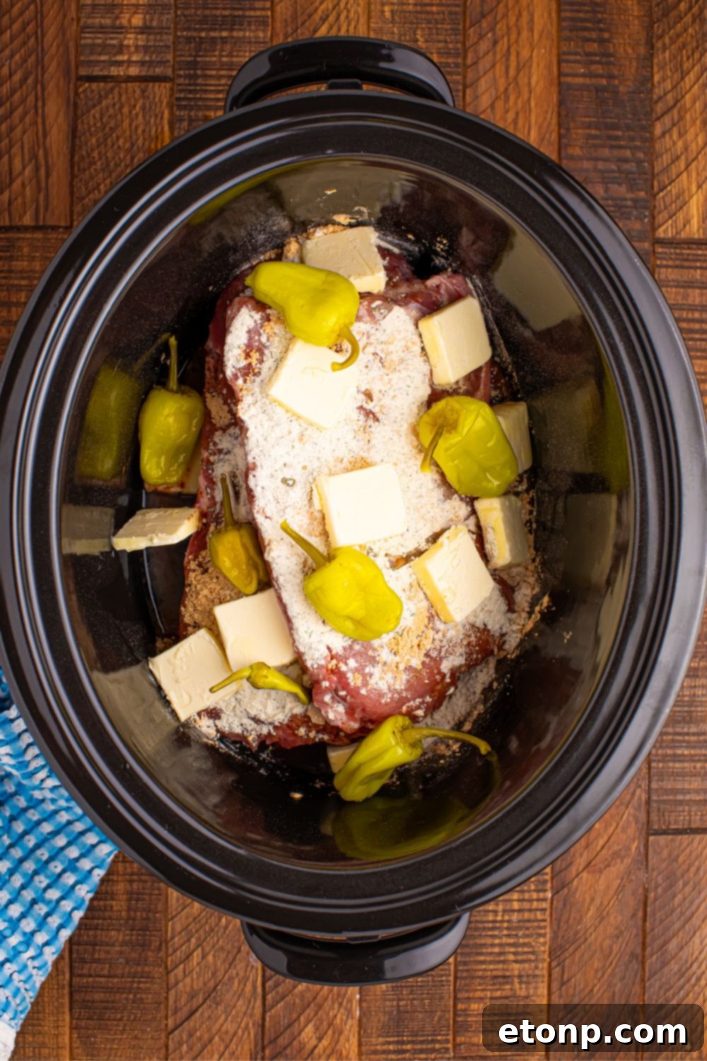 Raw ribs in a slow cooker, generously topped with ranch and au jus seasonings, whole pepperoncini peppers, and pats of unsalted butter.