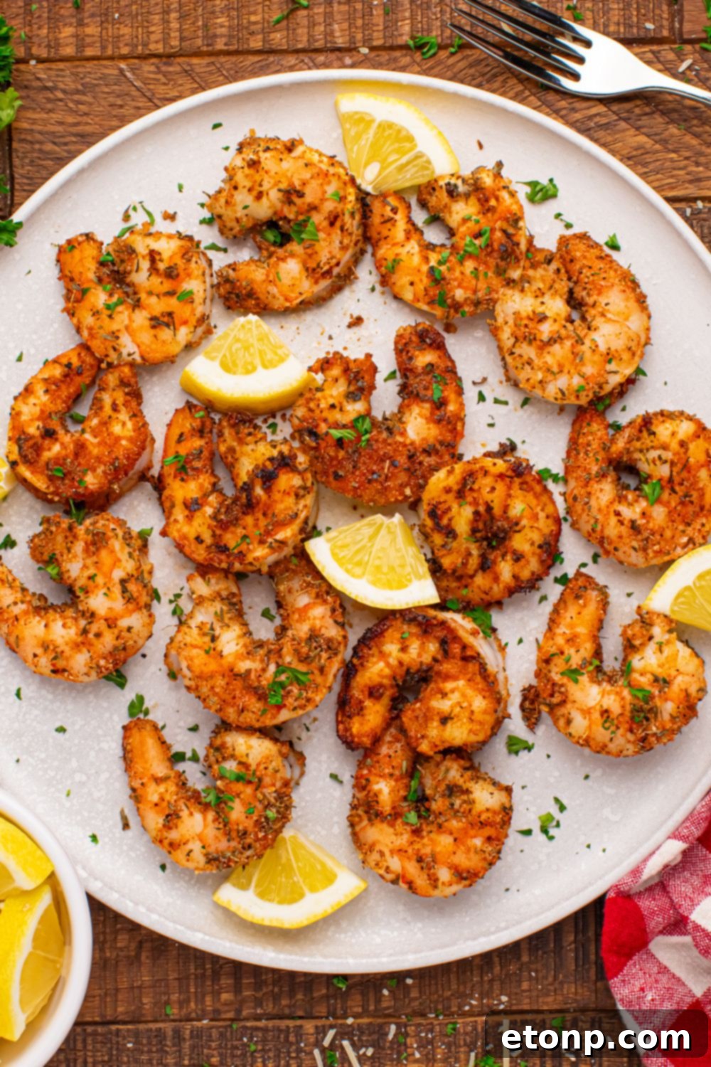 An overhead shot of a white plate generously scattered with perfectly grilled Cajun shrimp, fresh lemon wedges, and a sprig of parsley, presented on a light-colored wooden table.