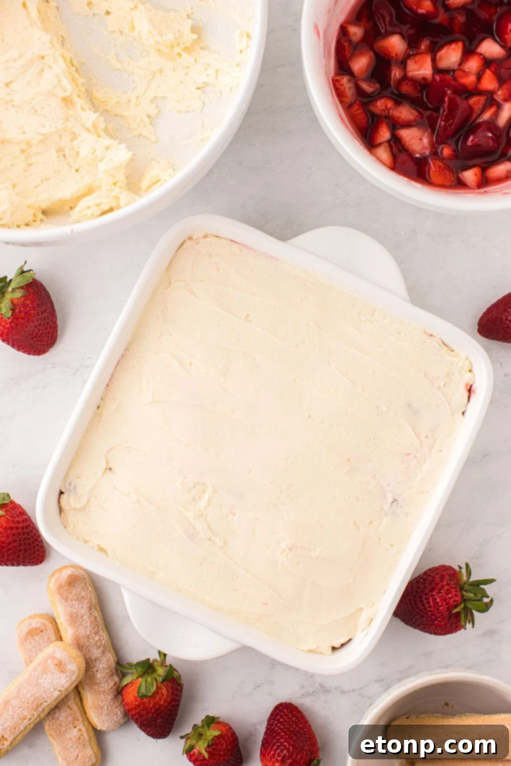 Strawberry Dream Tiramisu 9 The assembly process of Strawberry Tiramisu, showing layers being built in a pan, with a bowl of strawberries nearby.