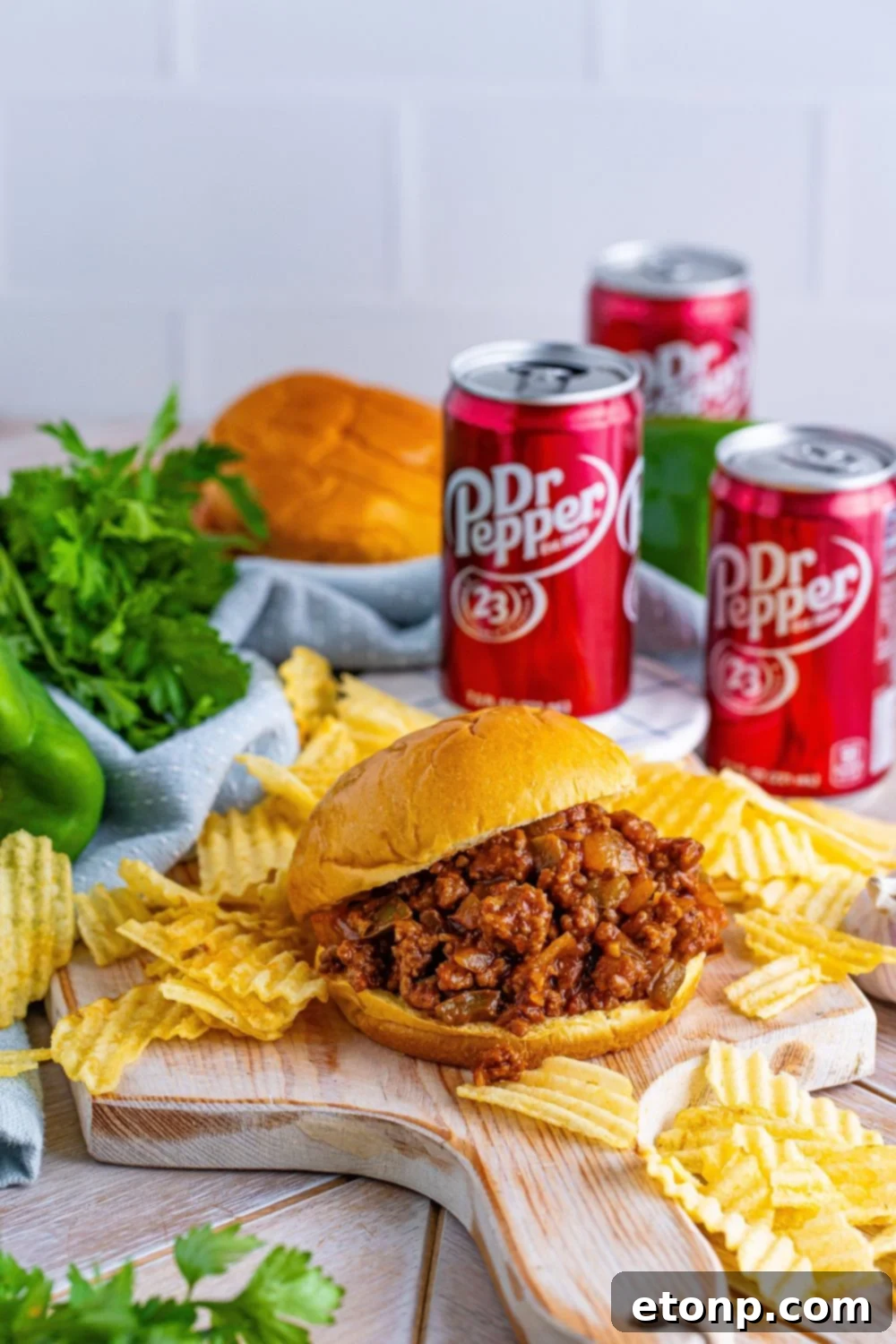 A delightful dinner spread featuring Dr. Pepper Sloppy Joes, a bowl of crispy chips, a can of Dr. Pepper, and fresh herbs scattered around a rustic table.