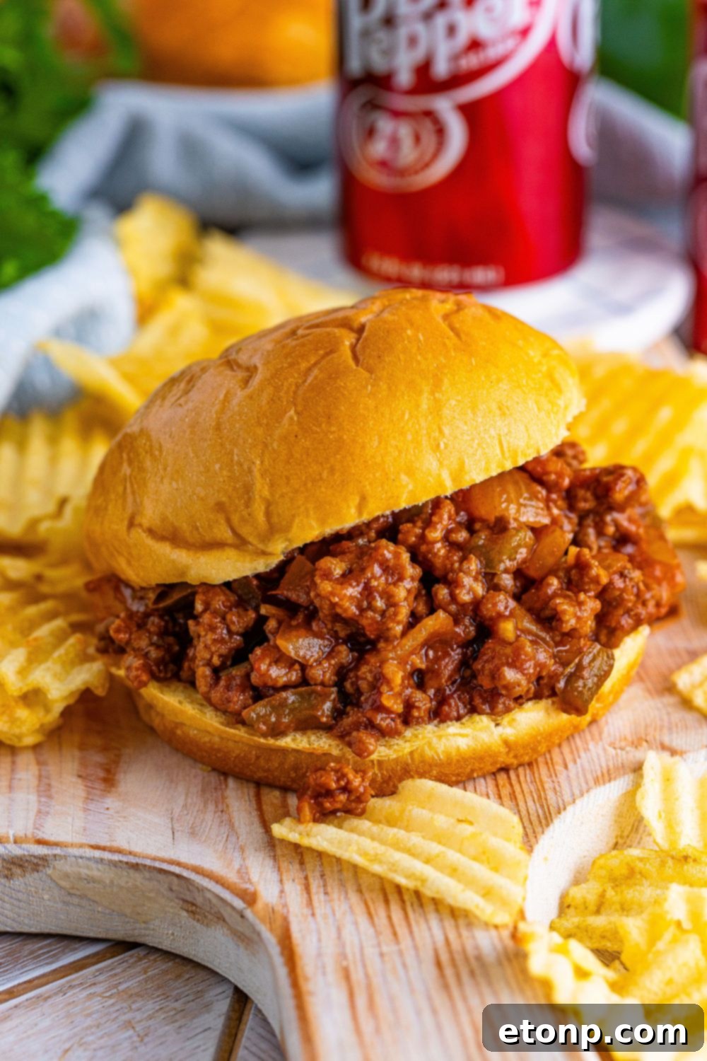 A Dr. Pepper Sloppy Joe sandwich on a cutting board, surrounded by plain potato chips and cans of soda, ready to be enjoyed.