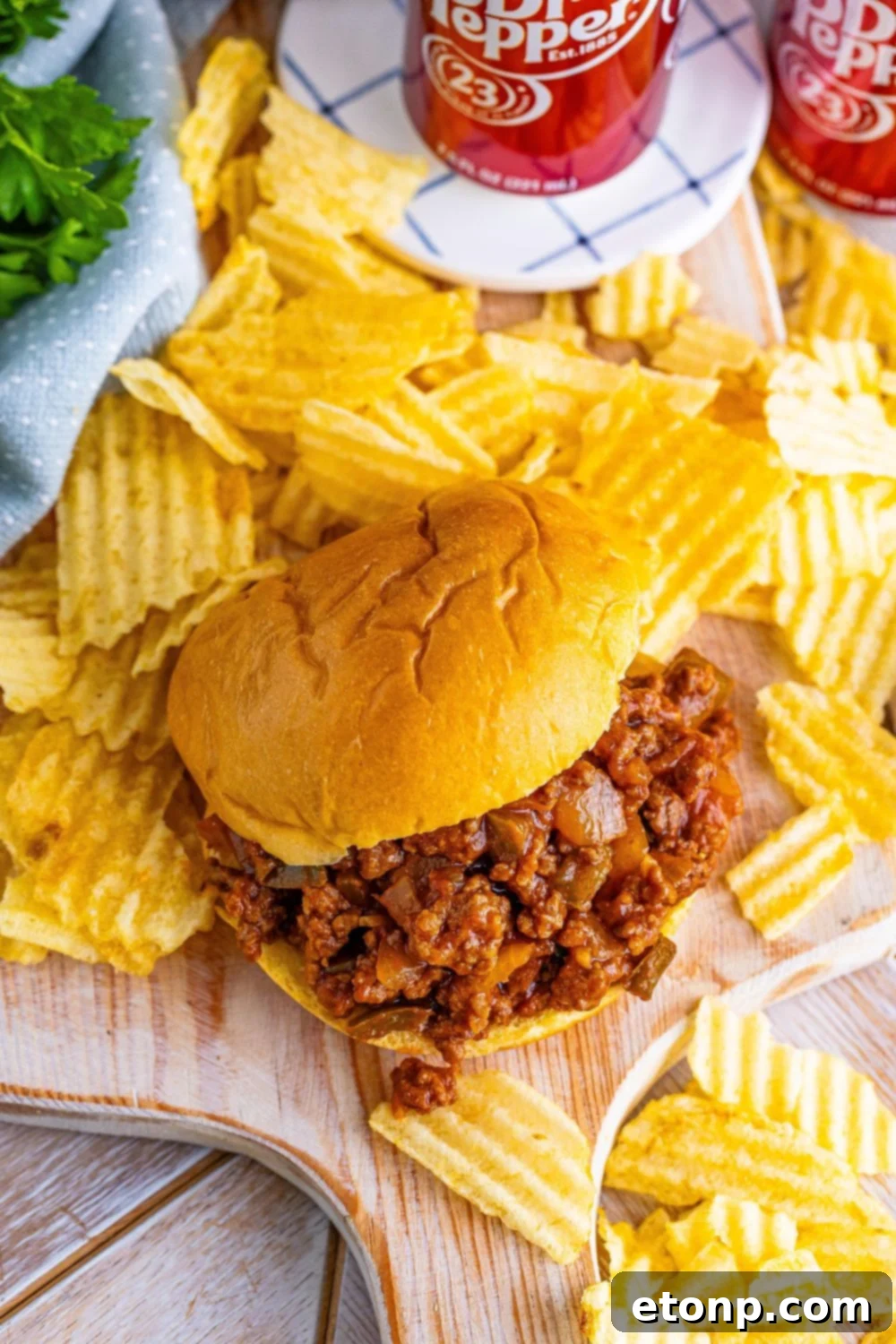 Three Dr. Pepper Sloppy Joes on a cutting board, garnished with fresh herbs, alongside potato chips and soda cans, creating a vibrant meal scene.
