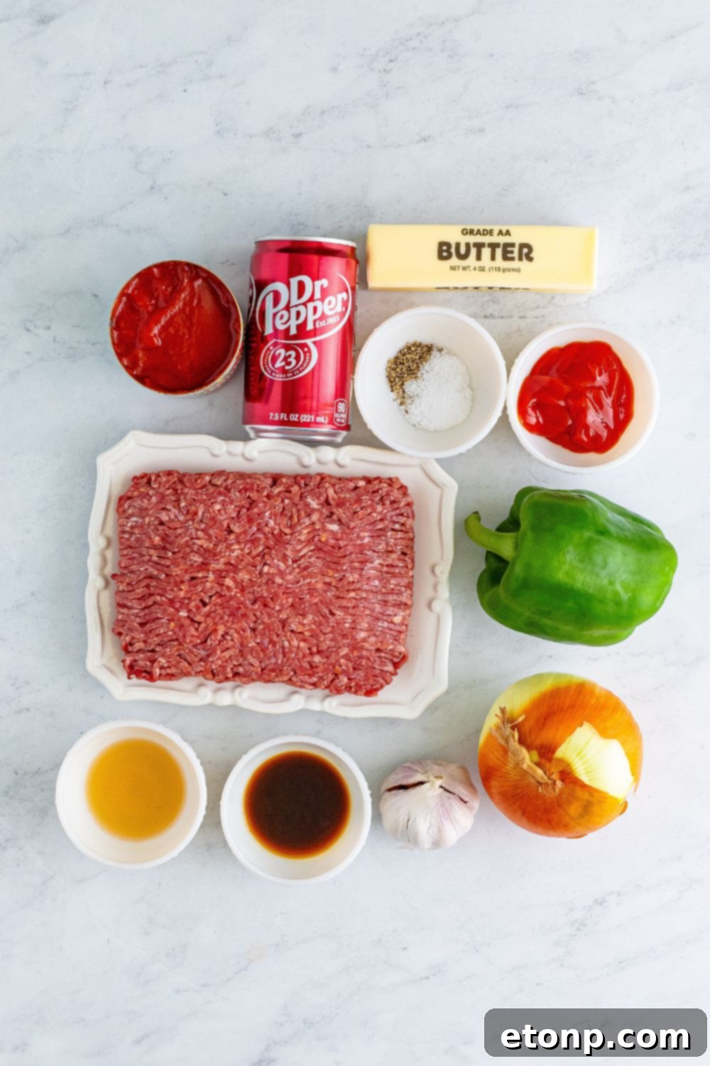 A vibrant flat lay of all the fresh ingredients for Dr. Pepper Sloppy Joes laid out on a marble countertop, including ground beef, vegetables, and condiments.