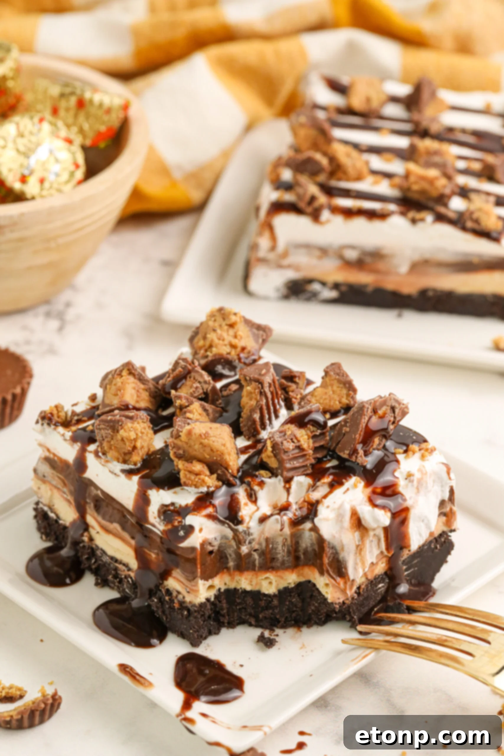 Reese's Peanut Butter Bliss Stack 2 A slice of Reese's Lasagna on a white plate with a bite taken out, showing the distinct layers of Oreo crust, peanut butter cream cheese, chocolate pudding, and whipped topping, adorned with crumbled Reese's Cups.