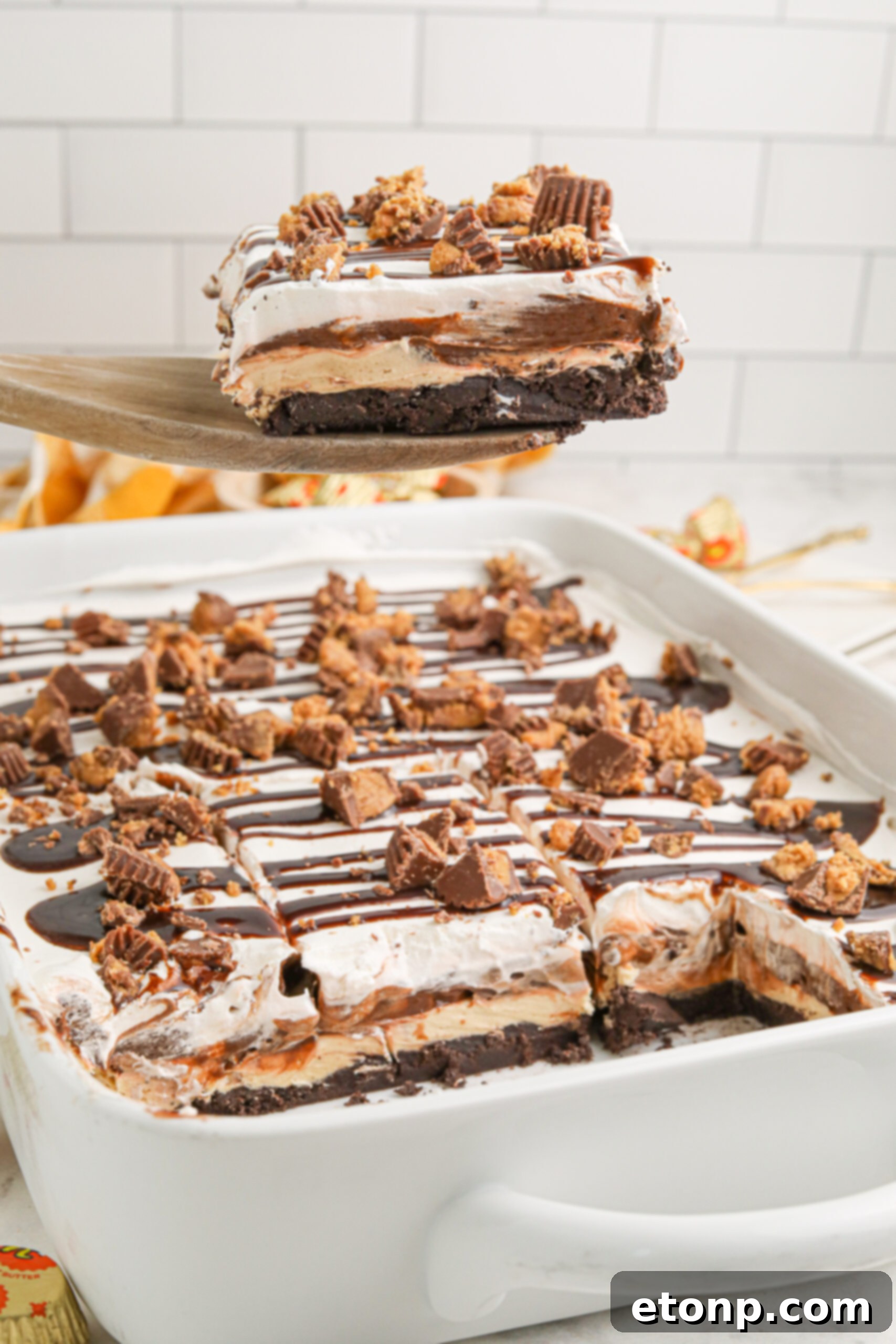 Reese's Peanut Butter Bliss Stack 17 Reese's Lasagna in a white baking pan, with a piece being carefully lifted, highlighting the texture and layers of the dessert.