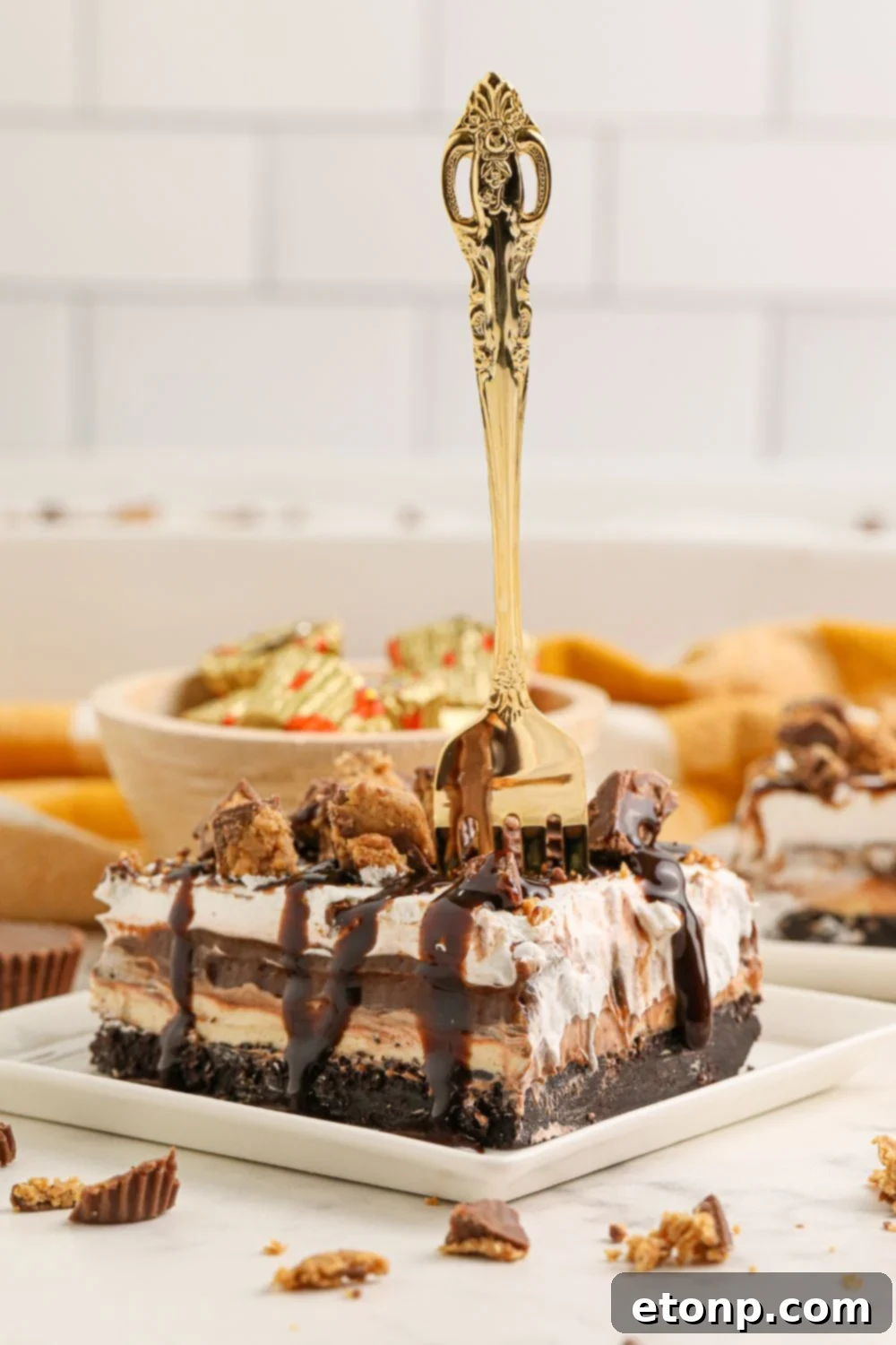 Reese's Peanut Butter Bliss Stack 4 A delectable slice of Reese's Lasagna on a table, complete with a fork, with a bowl of Reese's Peanut Butter Cups in the background, suggesting pure indulgence.