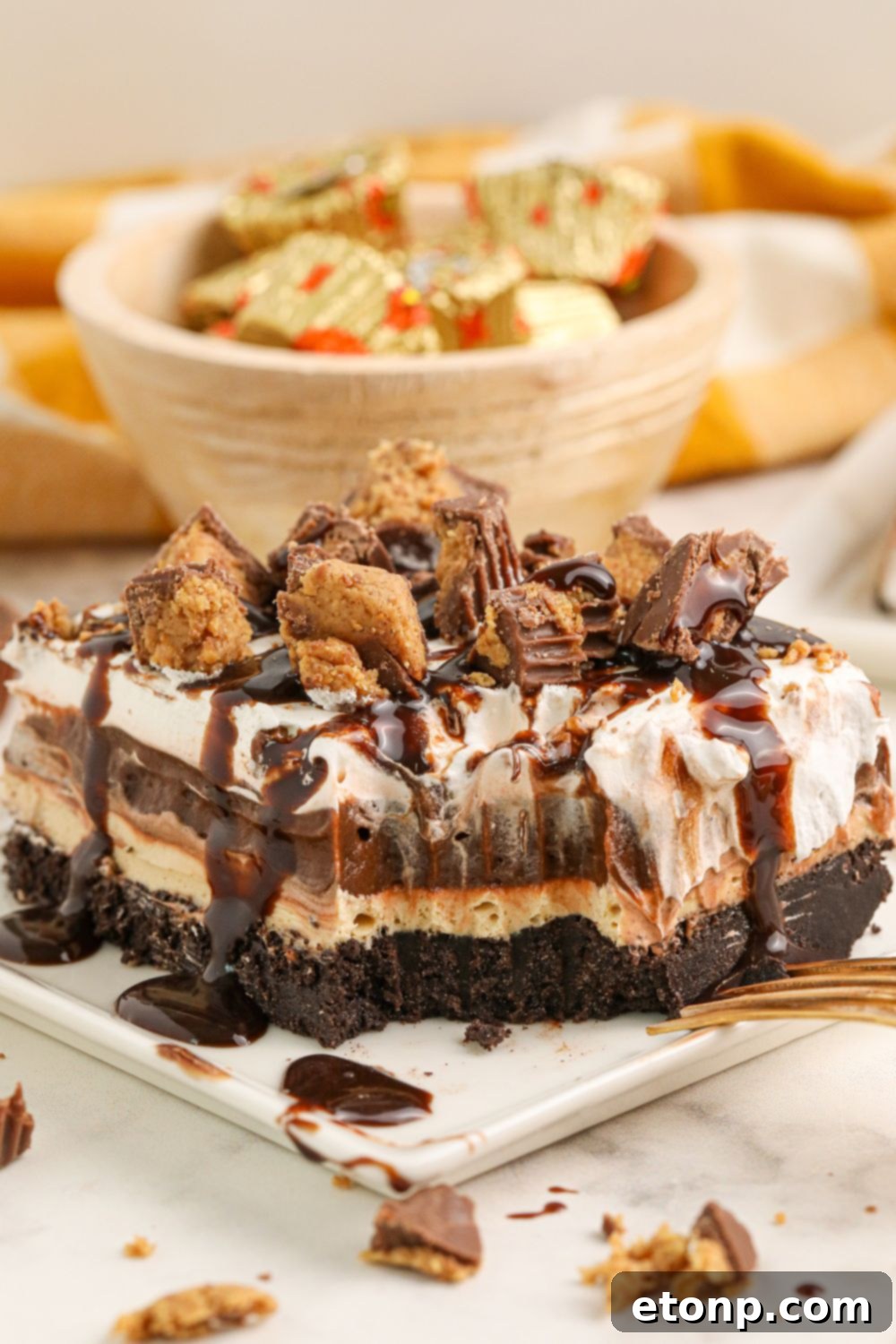 Reese's Peanut Butter Bliss Stack 5 A tantalizing slice of Reese's Lasagna on a plate with a bite removed, while a bowl of mini Reese's cups with wrappers rests enticingly in the background.