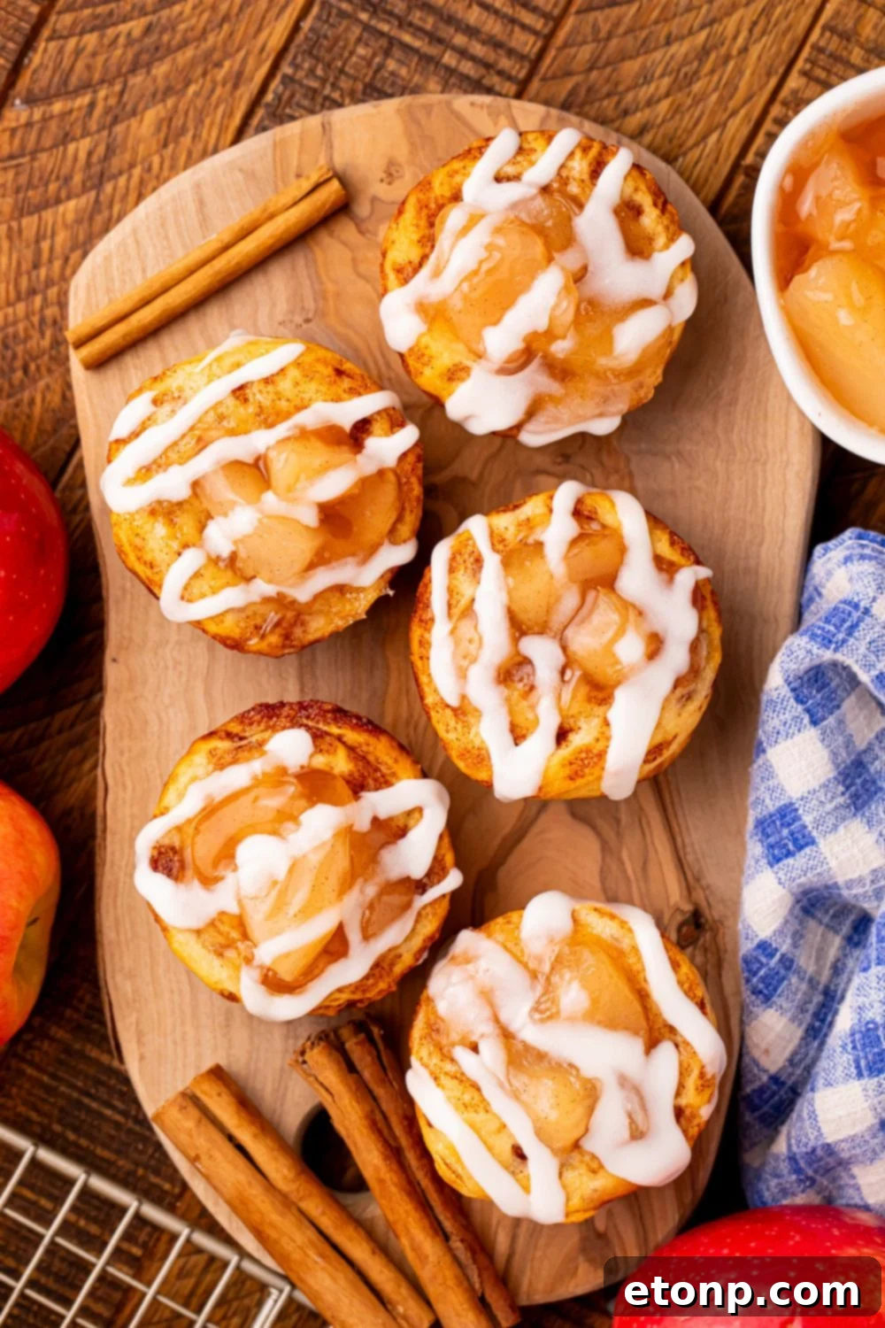 Effortless Apple Tartlets 14 apple pie cuts placed on a wooden tray with cinnamon sticks around the cups, and a bowl of pie filling and towel to the side