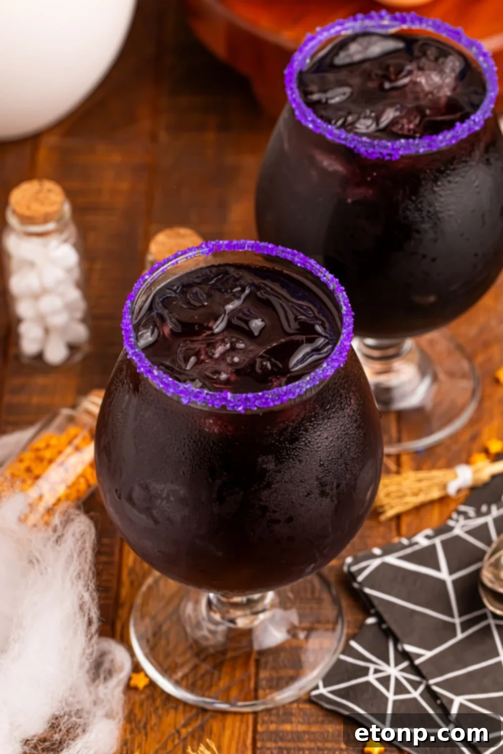 Witches Brew Cocktail on table with two glasses, a purple sugar rim and decor for Halloween all around it