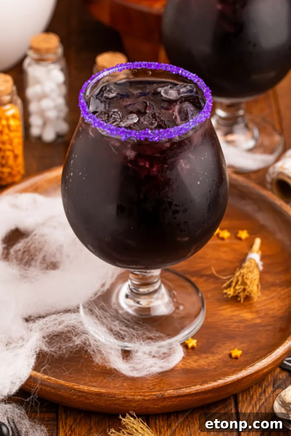 view of a glass with a deep purple cocktail in it with a light purple sugar rim sitting on table with Halloween decorations