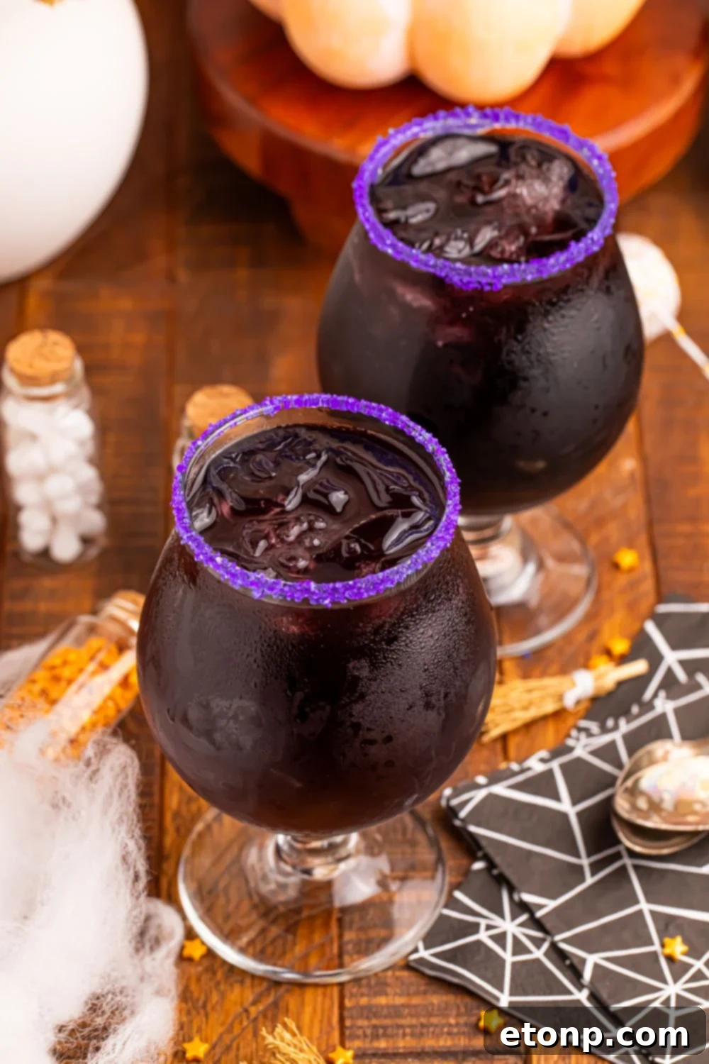 two glasses of witches brew cocktails sitting on wooden table with bottles of sprinkles in them, spider web napkins and more decor on table