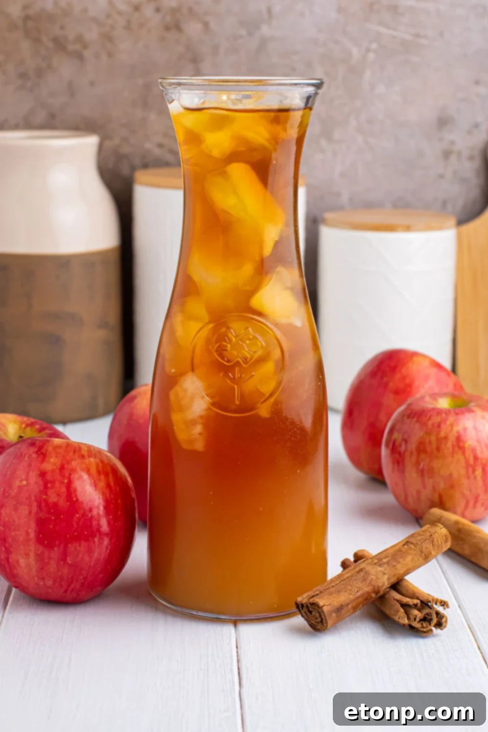 Orchard Apple Iced Tea 12 cider tea in a pitcher with cinnamon sticks and whole apples around it showing this is an apple cider iced tea