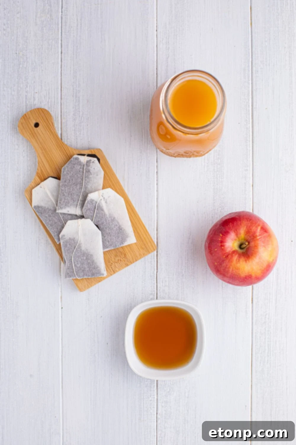 Orchard Apple Iced Tea 6 ingredients for making apple cider iced tea on a white table in single serve bowls: tea bags on a cutting board, mug of apple cider, whole apple