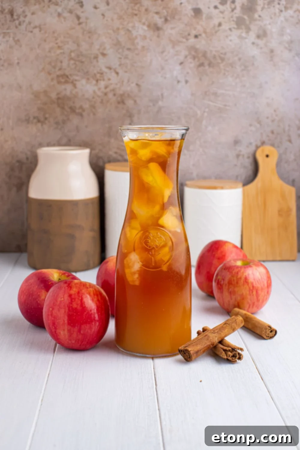 Orchard Apple Iced Tea 8 small pitcher of apple cider iced tea on table with apples and cinnamon sticks around it, and jars and cutting board in the background