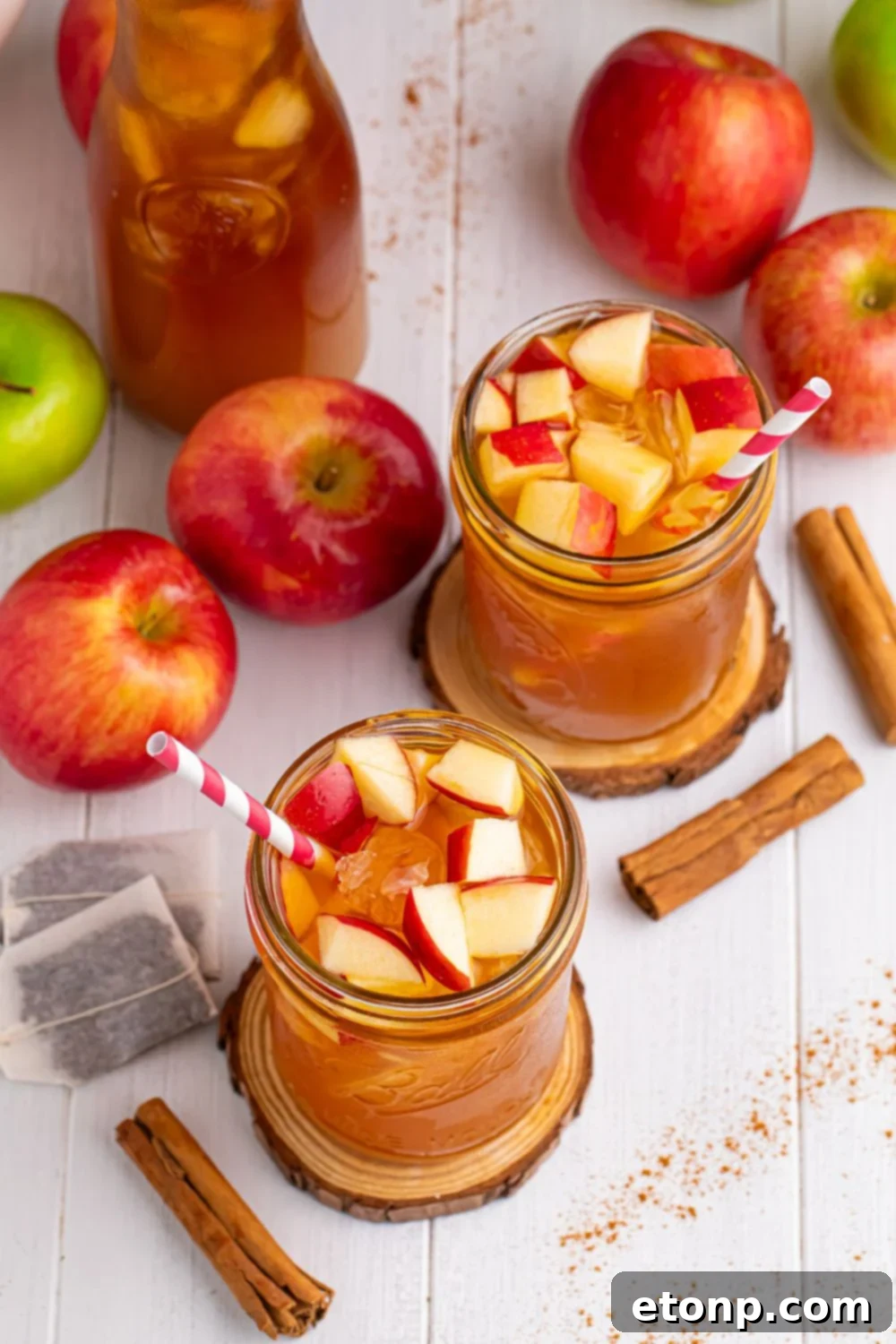 Orchard Apple Iced Tea 10 table setting with apple cider teas in glasses with straws. Tea bags, a variety of apples, cinnamon sticks and pitcher of tea all on table