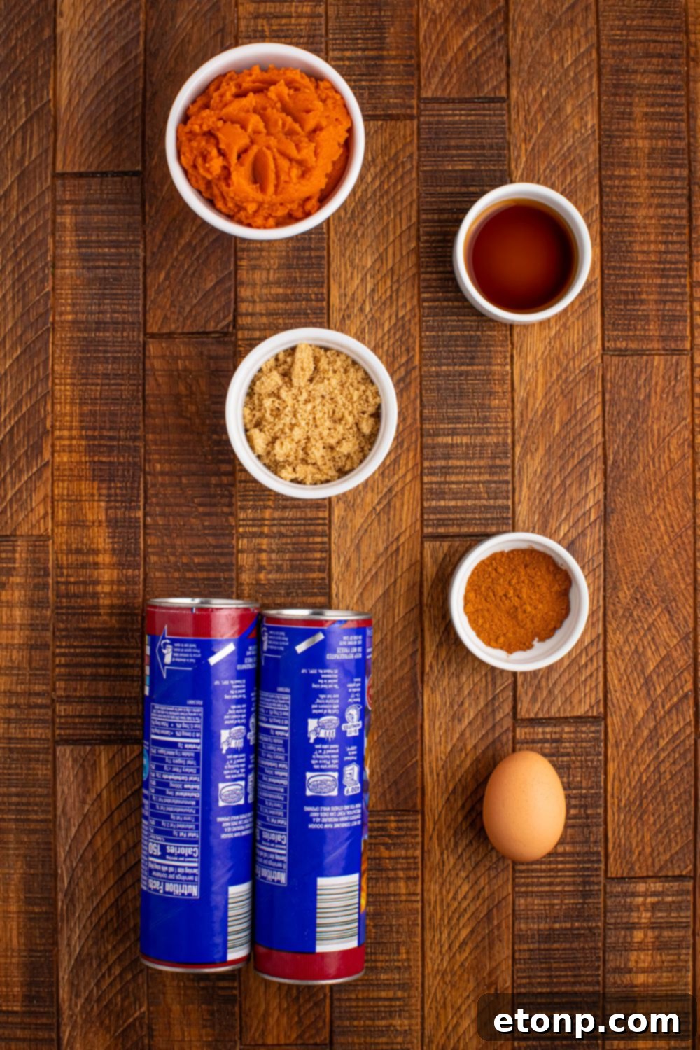 All the essential ingredients for Pumpkin Pie Cinnamon Roll Cups displayed on a table: canned cinnamon rolls, pumpkin puree, brown sugar, an egg, vanilla extract, and pumpkin pie spice.