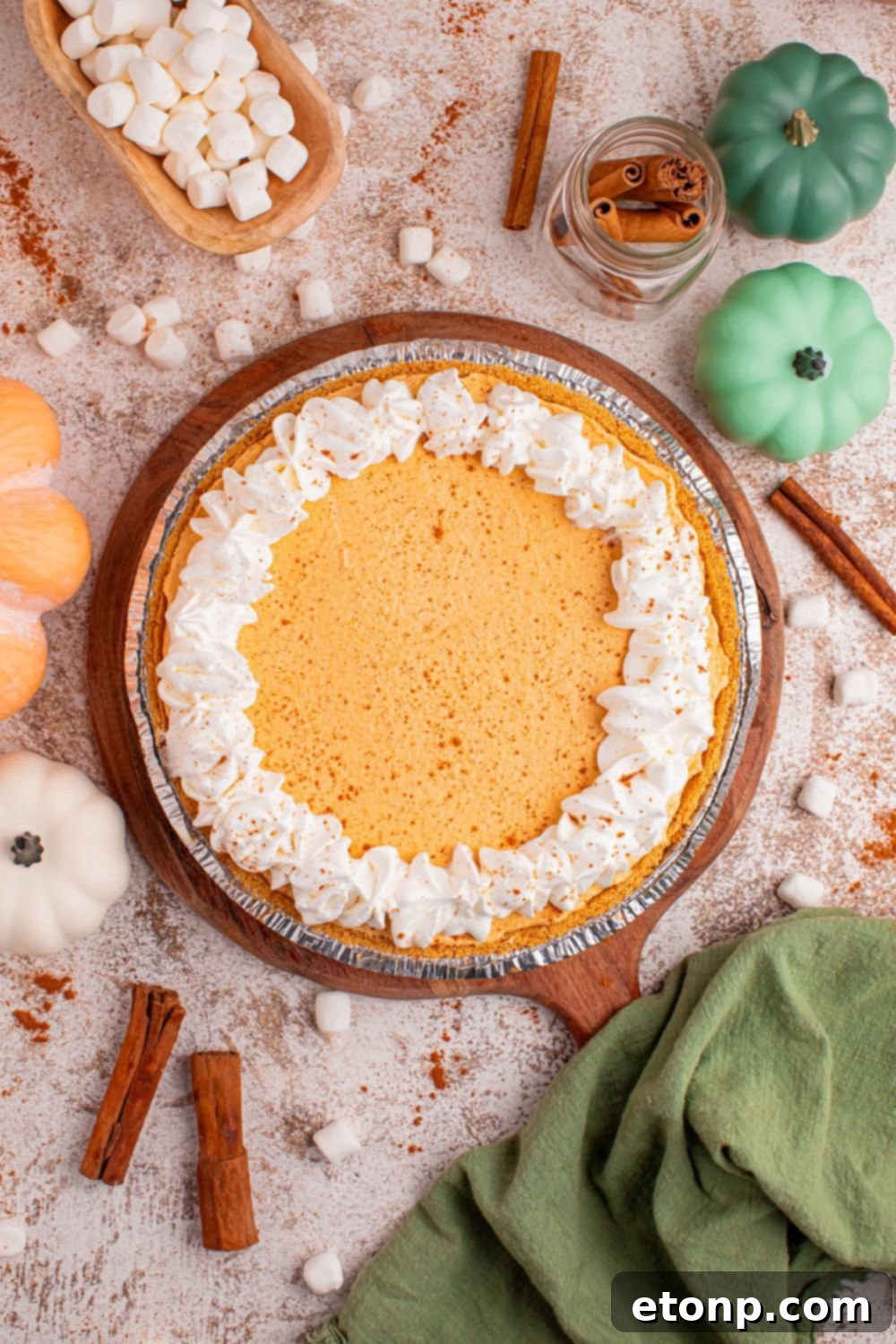 A beautifully presented Marshmallow Pumpkin Pie, lightly adorned with mini marshmallows, resting on a rustic wooden table amidst charming autumnal decorations and fragrant cinnamon sticks, creating a cozy fall ambiance.