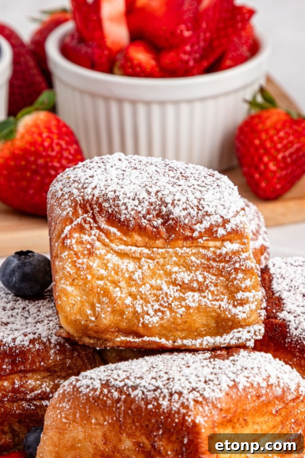 a close-up of Hawaiian Roll French Toast on a plate, with a bowl of fresh sliced strawberries in the background