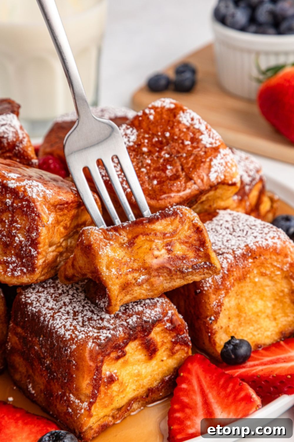 a fork lifting a bite of Hawaiian Roll French Toast from a plate adorned with sliced strawberries and blueberries