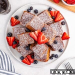 Hawaiian Roll French Toast stacked on a plate with powdered sugar and fruit on top of the French toast
