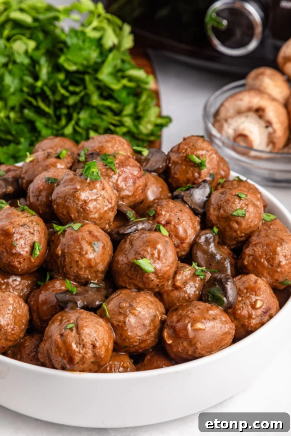 Comforting Slow Cooker Salisbury Steak Meatballs 15 white bowl with Salisbury Steak Meatballs in it and a bunch of parsley in a bundle behind bowl and mushrooms to the side