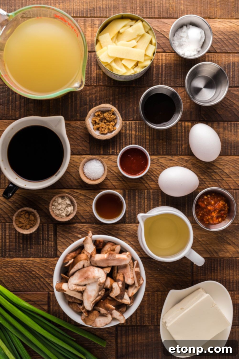 Various hot & sour soup ingredients displayed in small bowls on a wooden table, including mushrooms, tofu, and sauces
