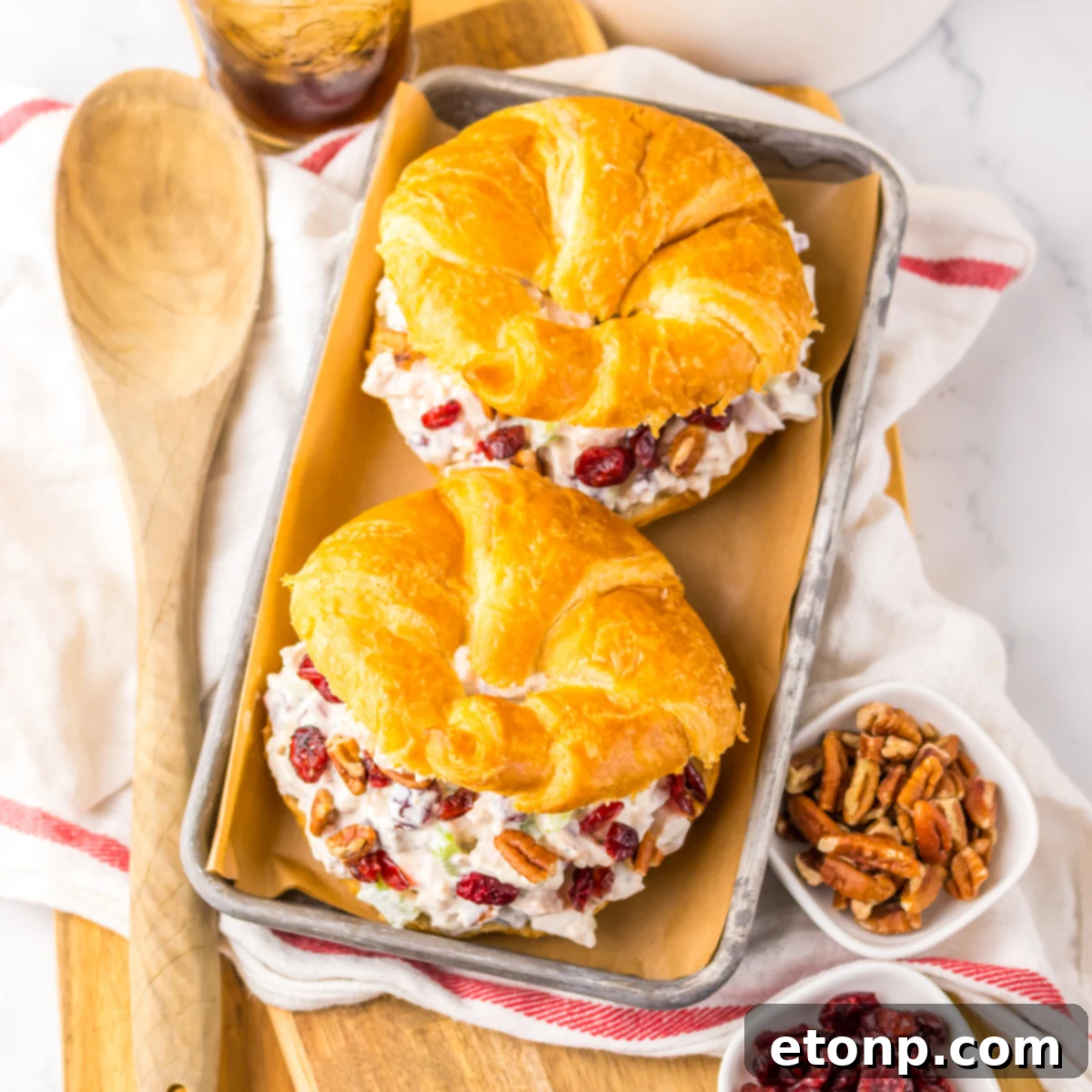 Close up view of a Cranberry Pecan Chicken Salad Croissant on a metal tray sitting on a red and white towel  with pecans and a wooden spoon to the side.  