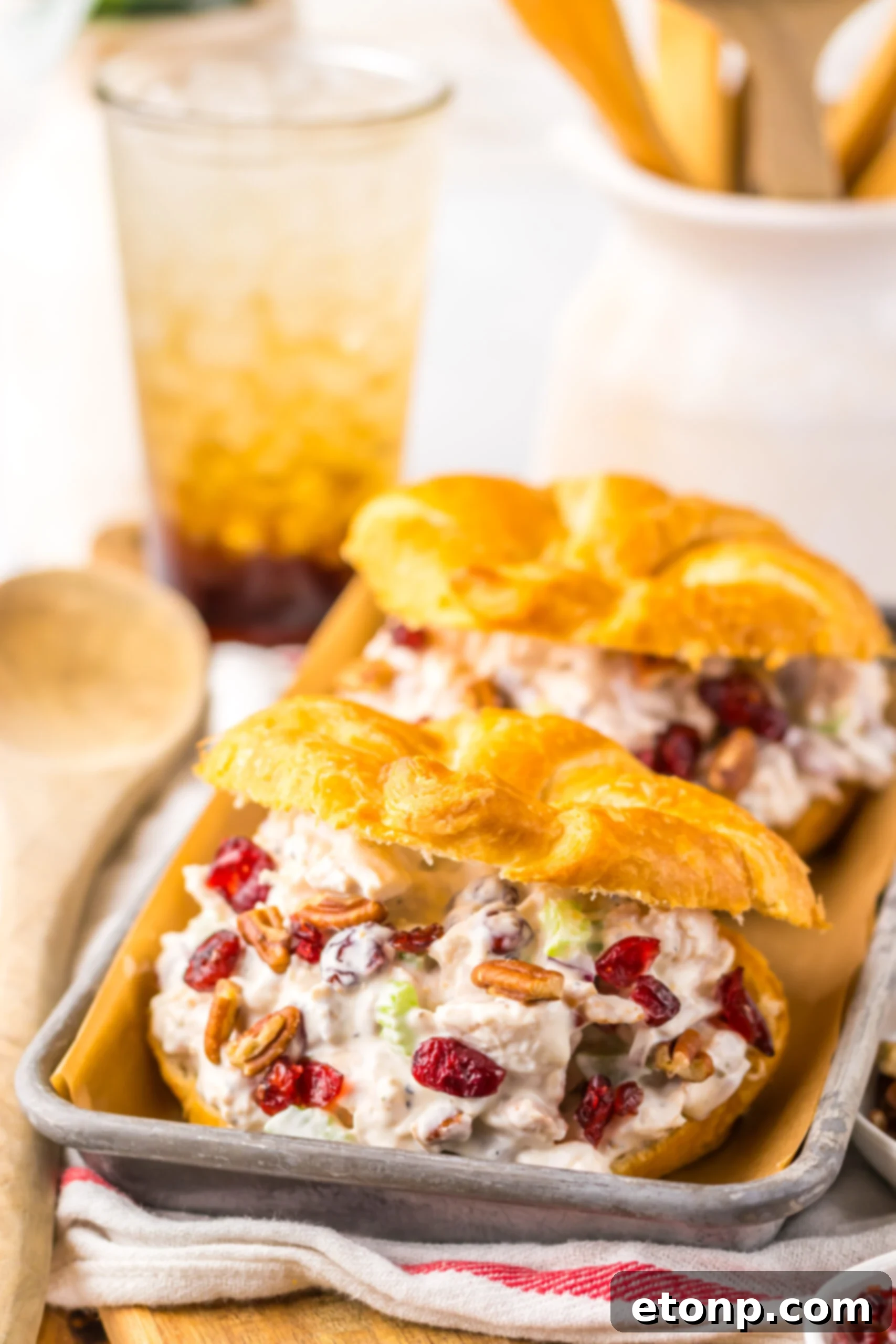 side view of a Cranberry Pecan Chicken Salad Croissant on a metal tray sitting with a glass of tea in the background