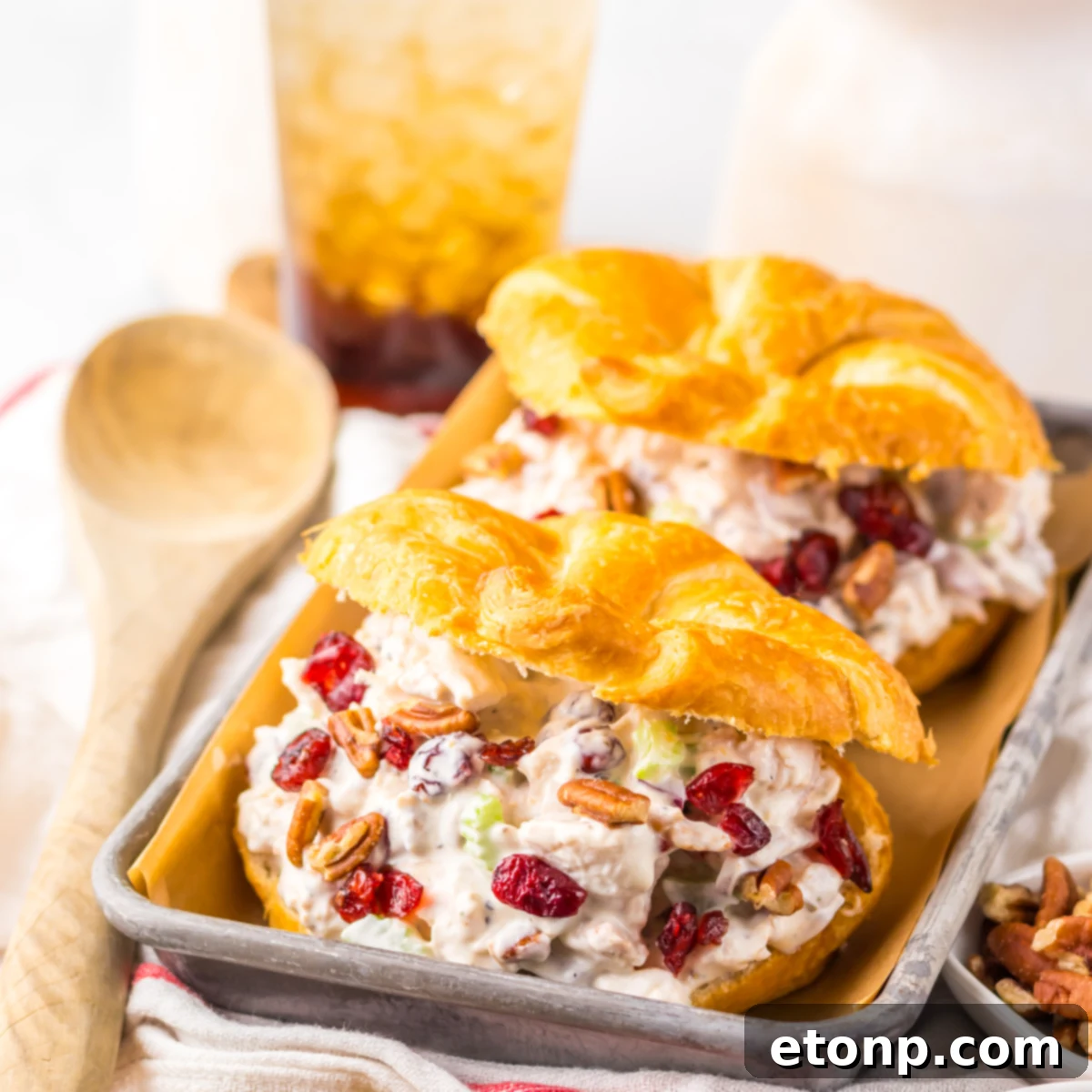 Close up view of a Cranberry Pecan Chicken Salad Croissant on a metal tray sitting on a red and white towel  with pecans and a wooden spoon tot he side.  