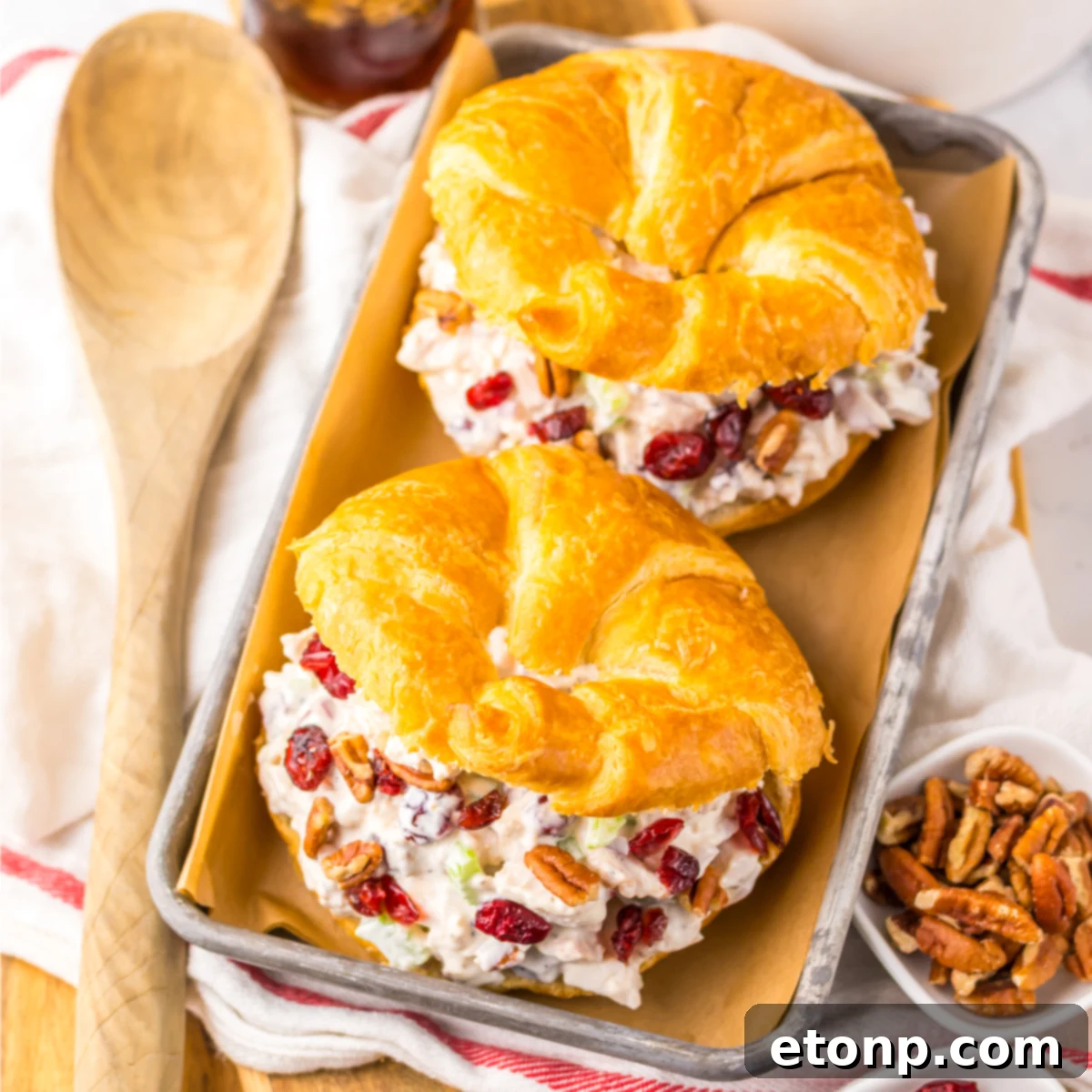 Over head view of a Cranberry Pecan Chicken Salad Croissant on a metal tray sitting on a red and white towel  with pecans and a wooden spoon to the side.  
