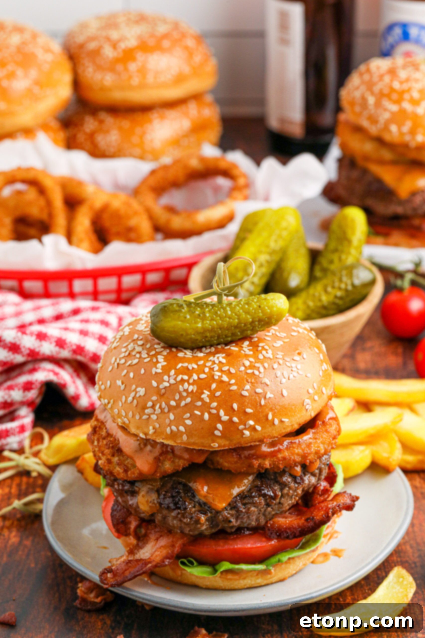 Cowboy Burger on a white plate that is fully loaded with toppings with a small bowl of pickles and fries in the background